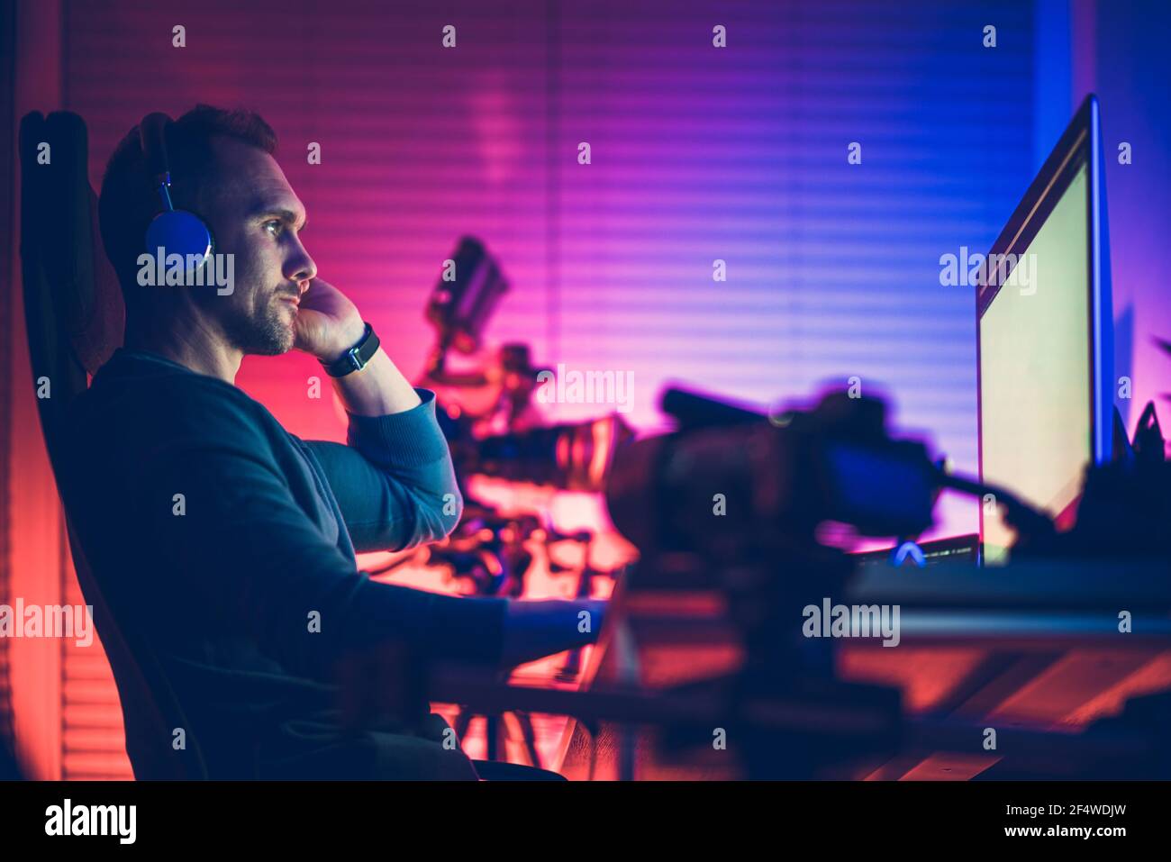 Kaukasische Männer in seinem 40s trägt Kopfhörer in seinem Home Office Bearbeitung Vlogs auf seinem Desktop-Computer. Kühle blaue und rote Raumbeleuchtung. Stockfoto