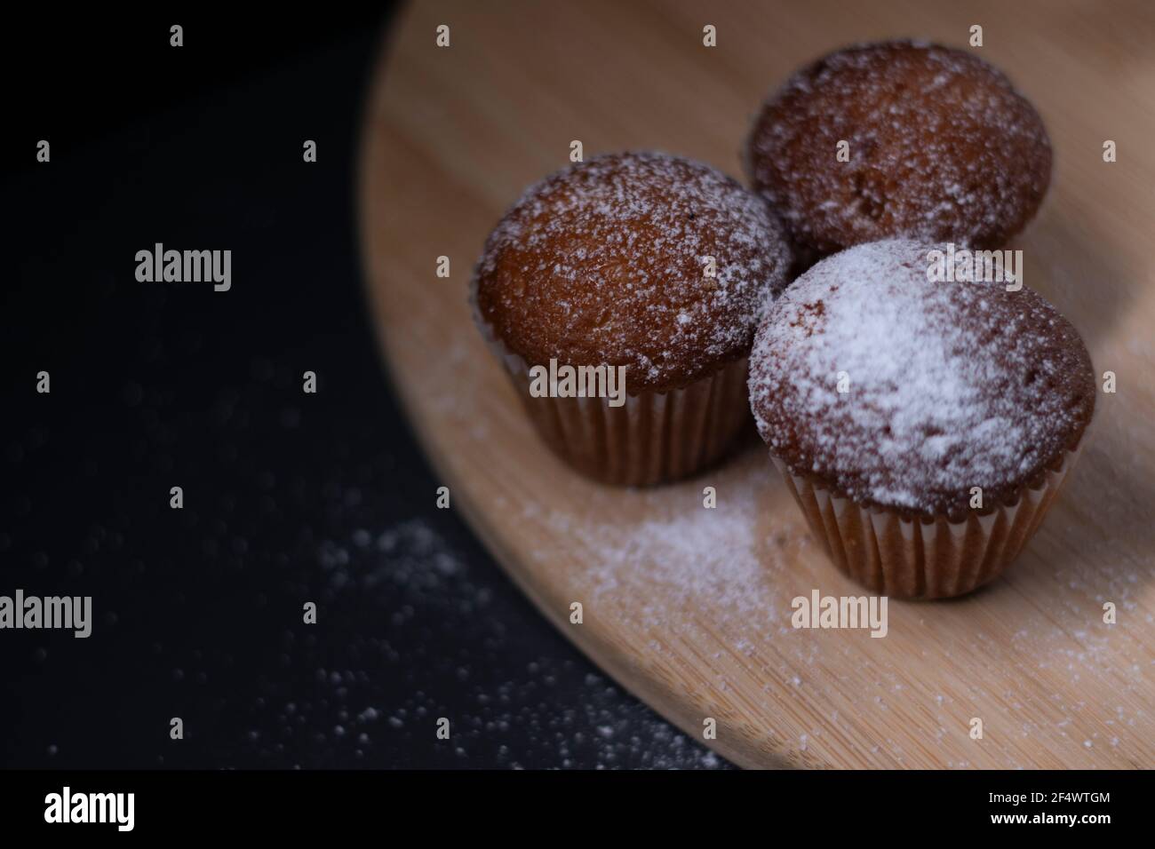 Drei Muffins mit Puderzucker bedeckt auf Holzständer isoliert auf schwarzem dunklen Hintergrund. Bäckerei süß Stockfoto