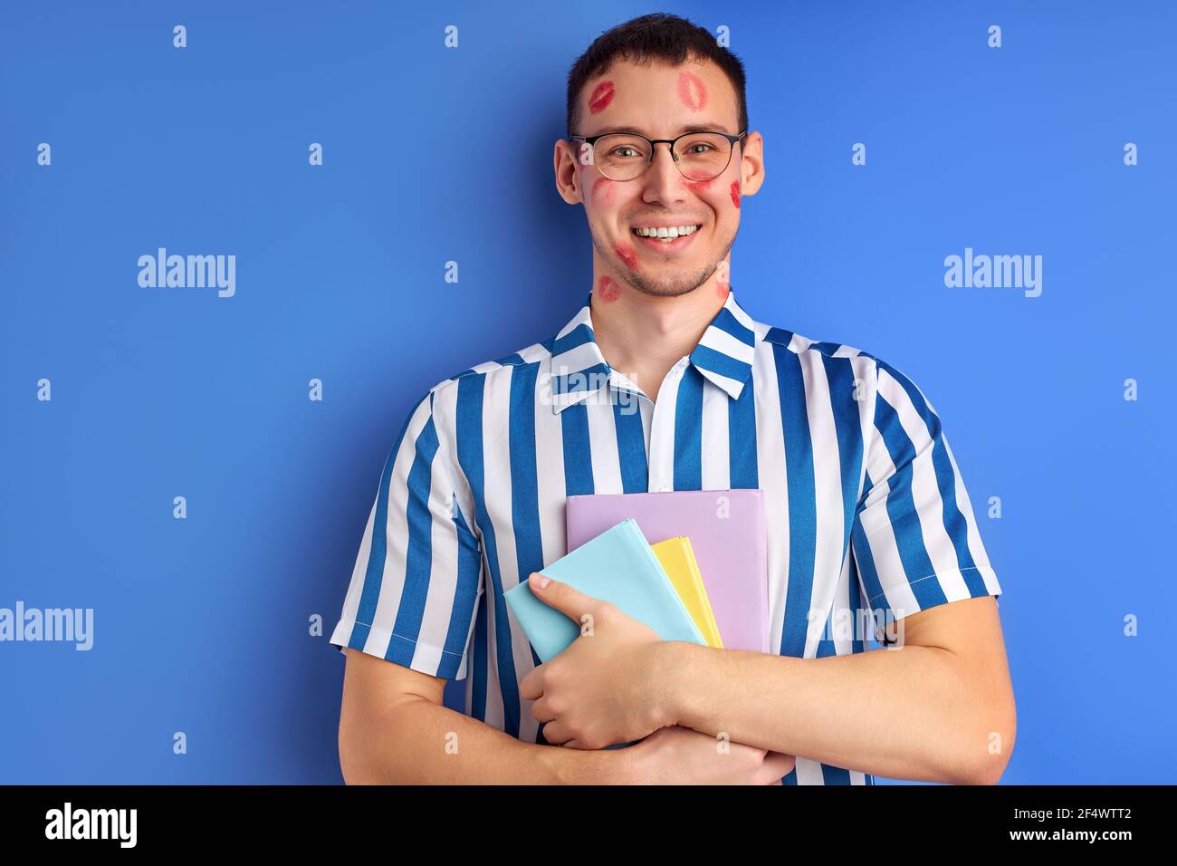 Glücklicher Student In Brillen Verliebt Sich In Erster Zeit Mit Kiss Abdrücke Auf Der Wange, Lächelnd An Der Kamera, Halten Bücher In Den Händen, Blauer Hintergrund Stockfoto Glücklicher Student In Brillen Verliebt Sich In Erster Zeit Mit Kiss Abdrücke Auf Der Wange, Lächelnd An Der Kamera, Halten Bücher In Den Händen, Blauer Hintergrund Stockfoto