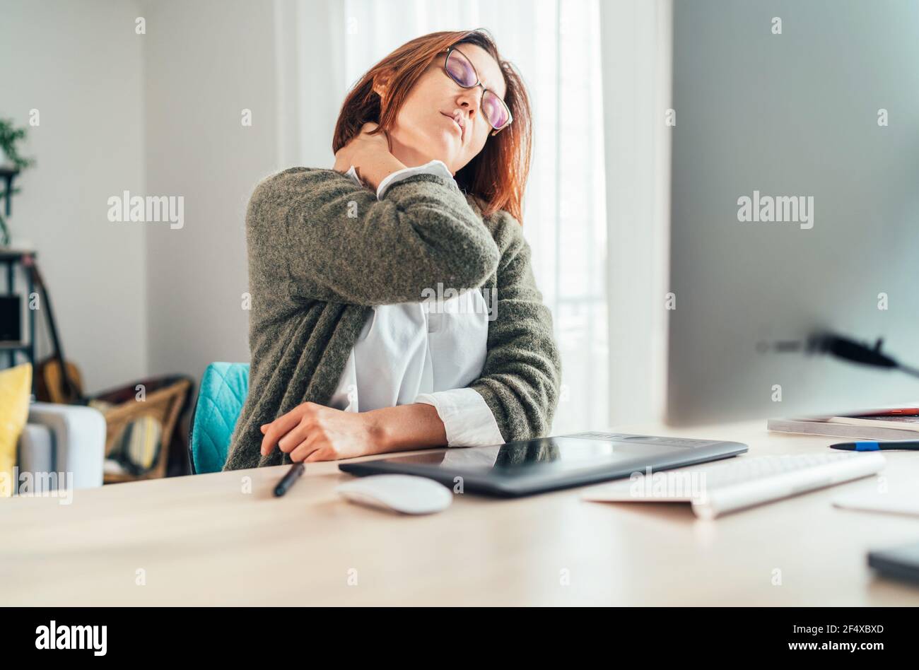Eine Frau mittleren Alters mit Nackenschmerzen, während sie als Grafikdesignerin mit einem modernen Computer in seinem Wohnzimmer arbeitet. Fernarbeit oder Freiberufler an w Stockfoto