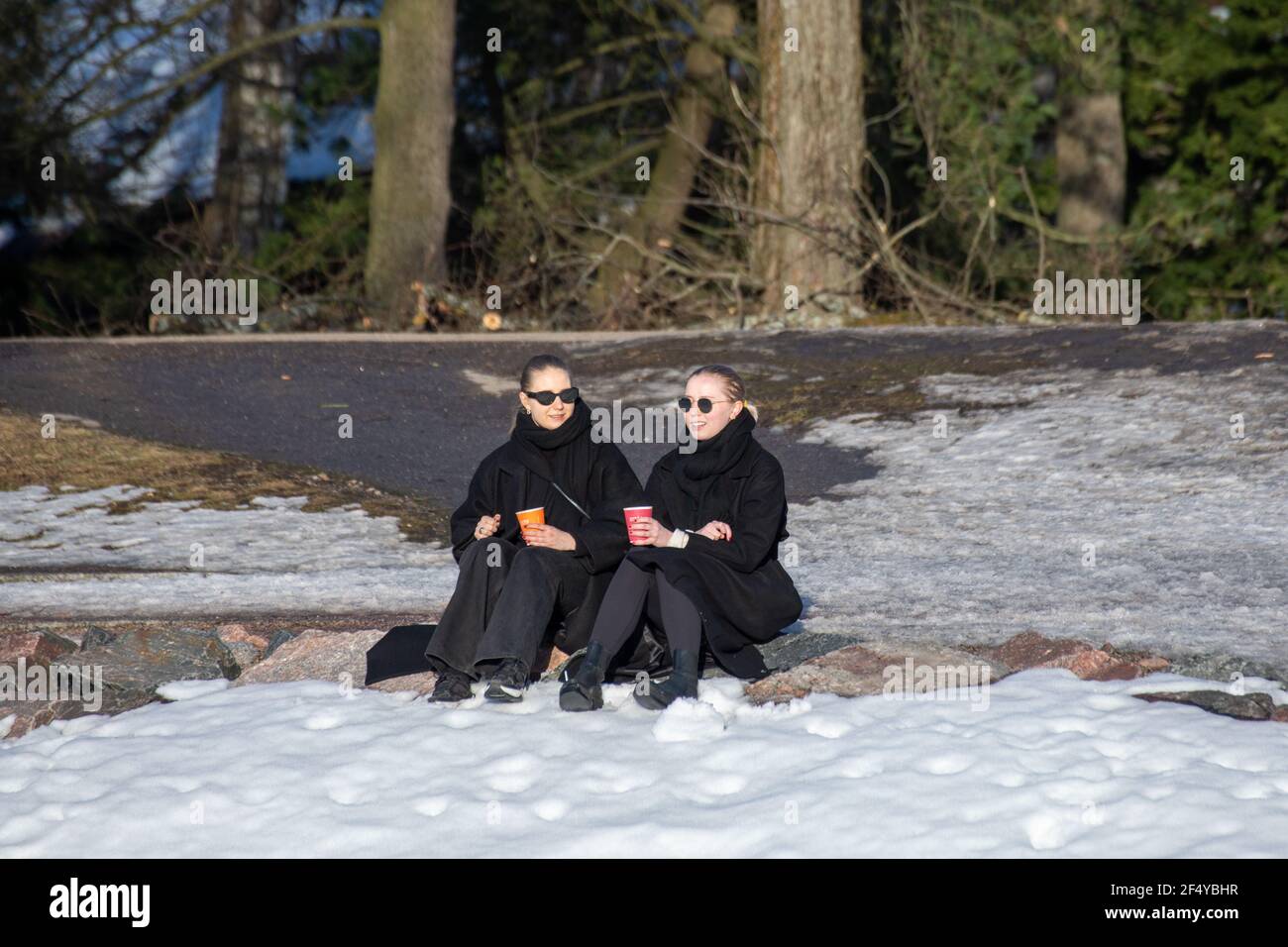 Junge Frauen genießen Getränke auf einem Uferstein während der Restaurantsperrung am frühen Frühlingsnachmittag im Stadtteil Taka-Töölöö in Helsinki, Finnland Stockfoto