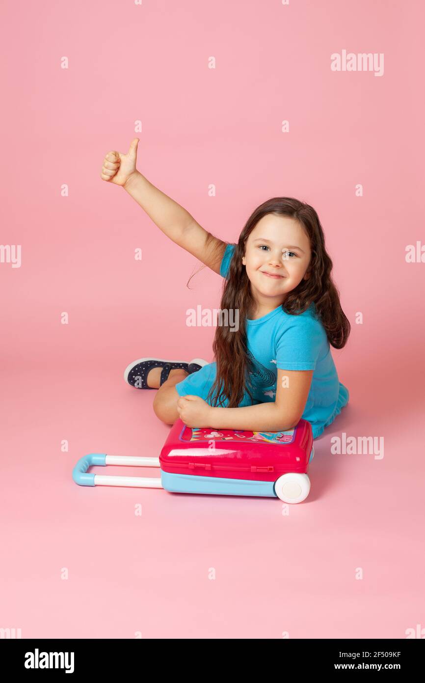Mädchen auf dem Boden mit ihren Händen auf einem karmesinroten Plastikkoffer und Daumen nach oben auf ihrem ausgestreckten Arm, isoliert auf einem rosa Hintergrund Stockfoto