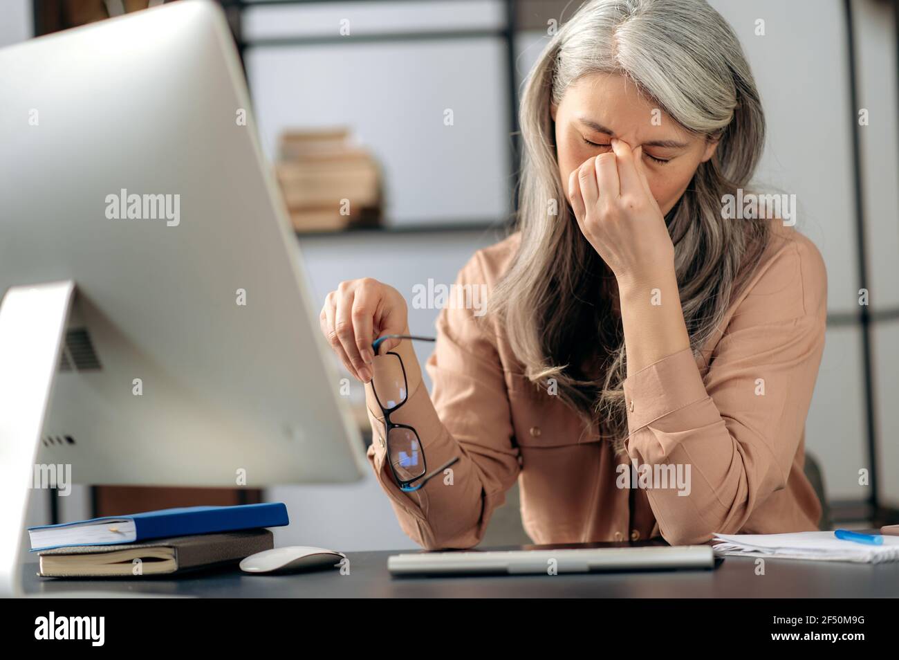 Überfordert müde traurige ältere grauhaarige Asiatin, Freiberuflerin, Managerin, hält Brille in der Hand, in stilvoller Kleidung, Kopfschmerzen, Stress nach langer Computerarbeit, massieren der Augen am Arbeitsplatz Stockfoto