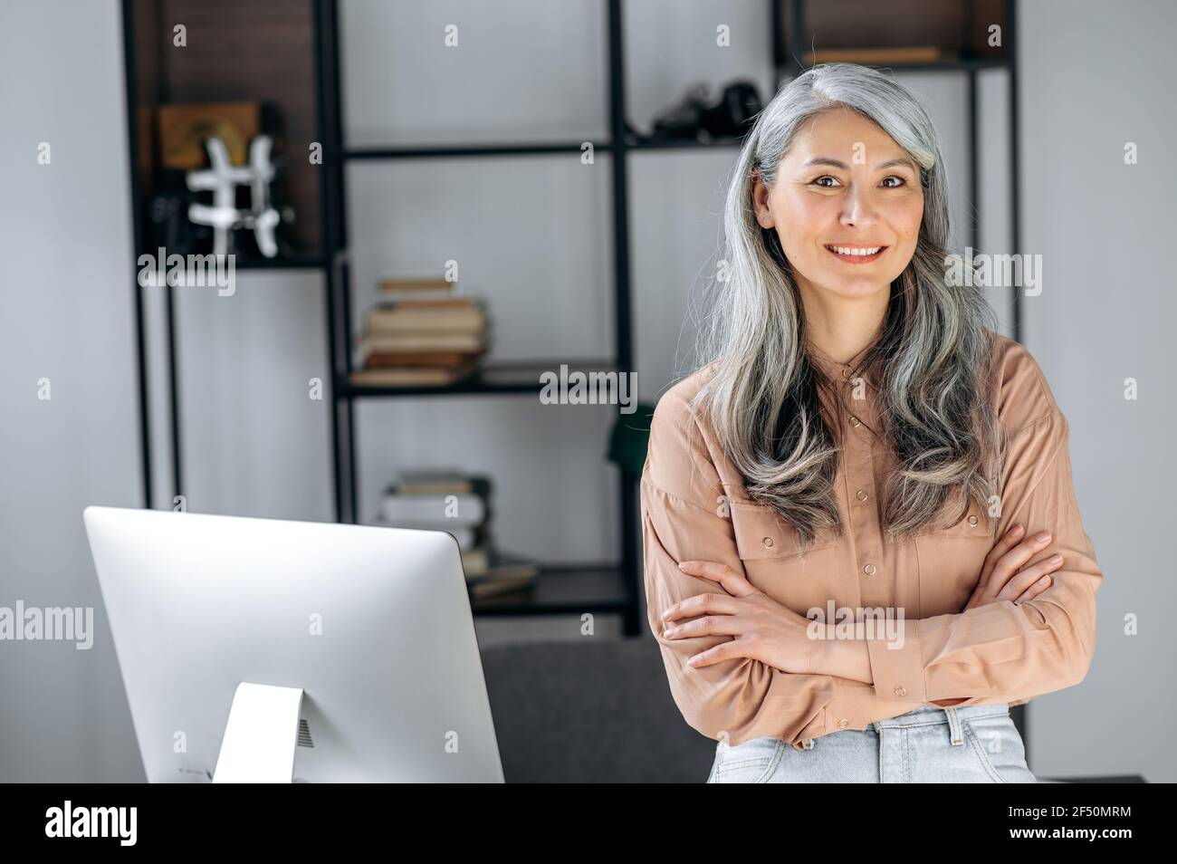 Porträt einer schönen erfolgreichen selbstbewussten grauhaarigen asiatischen Frau, Geschäftsfrau, Top-Managerin, steht in einem modernen Büro mit gekreuzten Armen, in stilvoller Kleidung, blickt auf die Kamera und lächelt freundlich Stockfoto