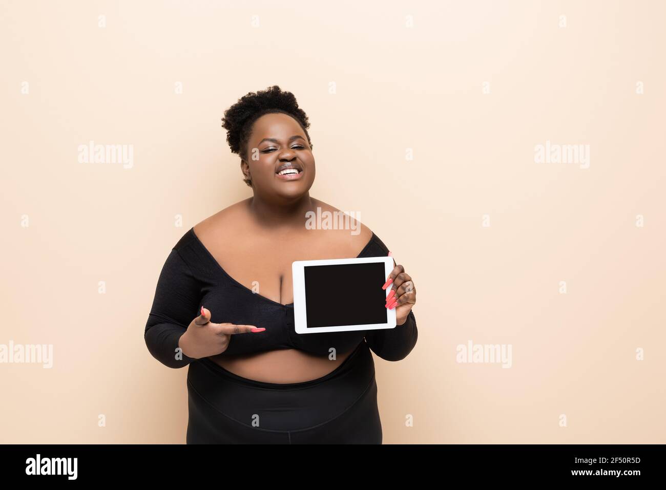 Happy african american plus size Frau zeigt mit dem Finger auf Digitales Tablet mit leerem Bildschirm isoliert auf beige Stockfoto