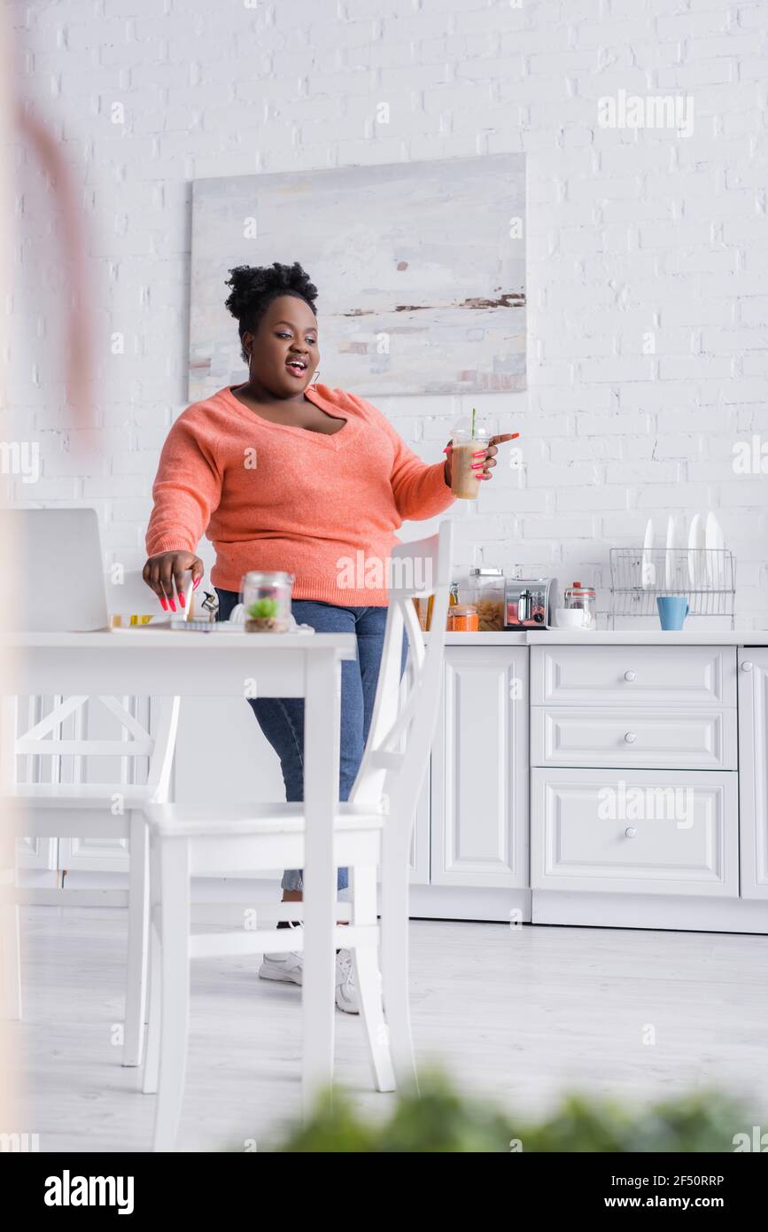 Happy african american plus size Frau hält Kunststoff-Tasse mit Smoothie in der Küche mit verschwommenem Vordergrund Stockfoto