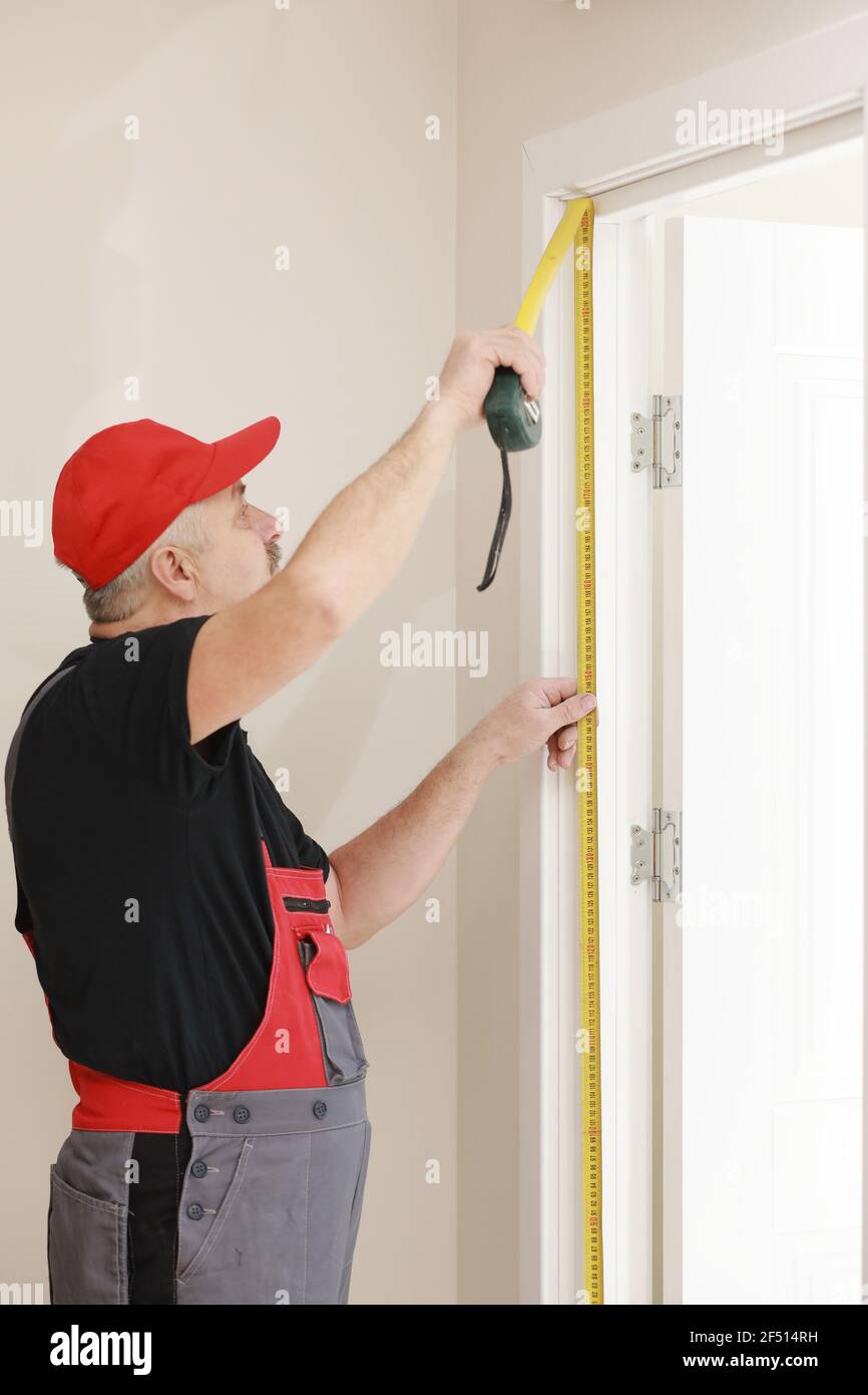 Arbeiter in roter Kappe und Arbeitsanzug mit dem Maßband. Man installiert die Türen. Messen Sie das Band in den Händen. Reparaturarbeiten. Wartung in den Wohnungen Stockfoto