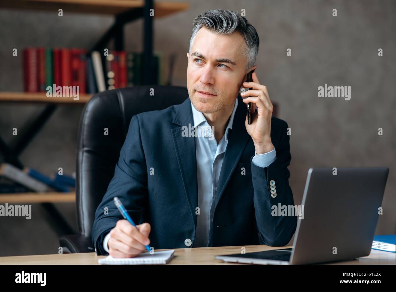 Selbstbewusst erfolgreich intelligente kaukasischen reifen Immobilienmakler, sitzen im Büro, in stilvollen Anzug gekleidet, kommuniziert mit einem Kunden durch ein Handy, macht Notizen, sieht zur Seite Stockfoto