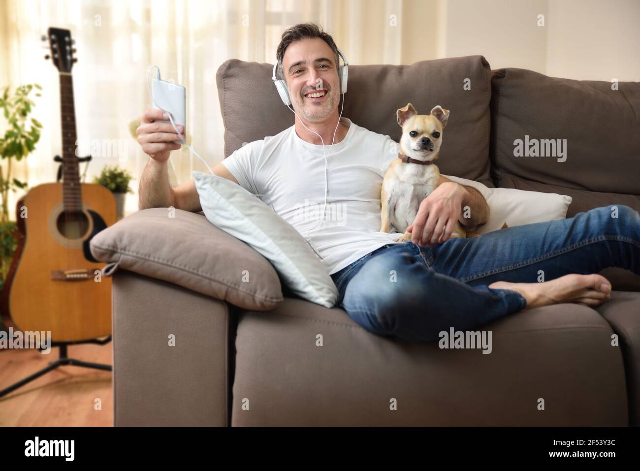 Entspannter Mann zu Hause auf dem Sofa liegend und lauscht Musik von einem Handy mit weißen Kopfhörern, die auf seine chihuahua Hund in seinen Armen Stockfoto