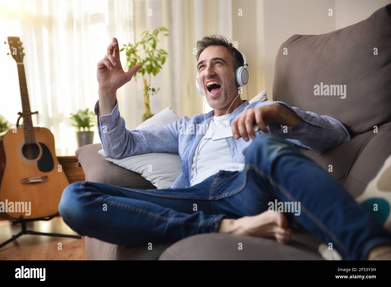 Euphorischer erwachsener Mann, der auf dem Sofa tanzt und Musik hört Von einem Handy mit weißen Kopfhörern in der lässigen gekleidet Wohnzimmer mit Fenster und Klimaanlage Stockfoto