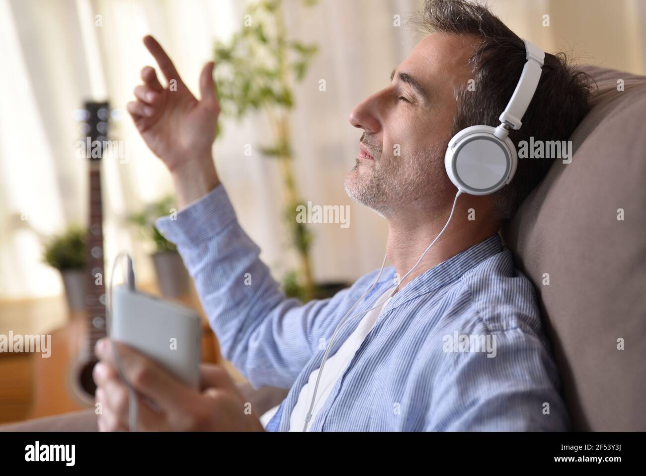 Detail des Mannes sitzt auf dem Sofa zu Hause hören Musik von einem Mobiltelefon mit weißen Kopfhörern Eintritt in ein warmes Sonnenlicht durch Th Stockfoto