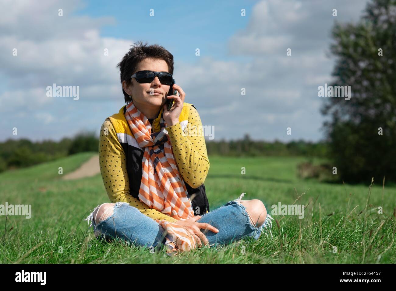 Frau mit Sonnenbrille, gelbem Mantel und Jeans sitzt auf der Wiese an sonnigen warmen Tag im Park Stockfoto