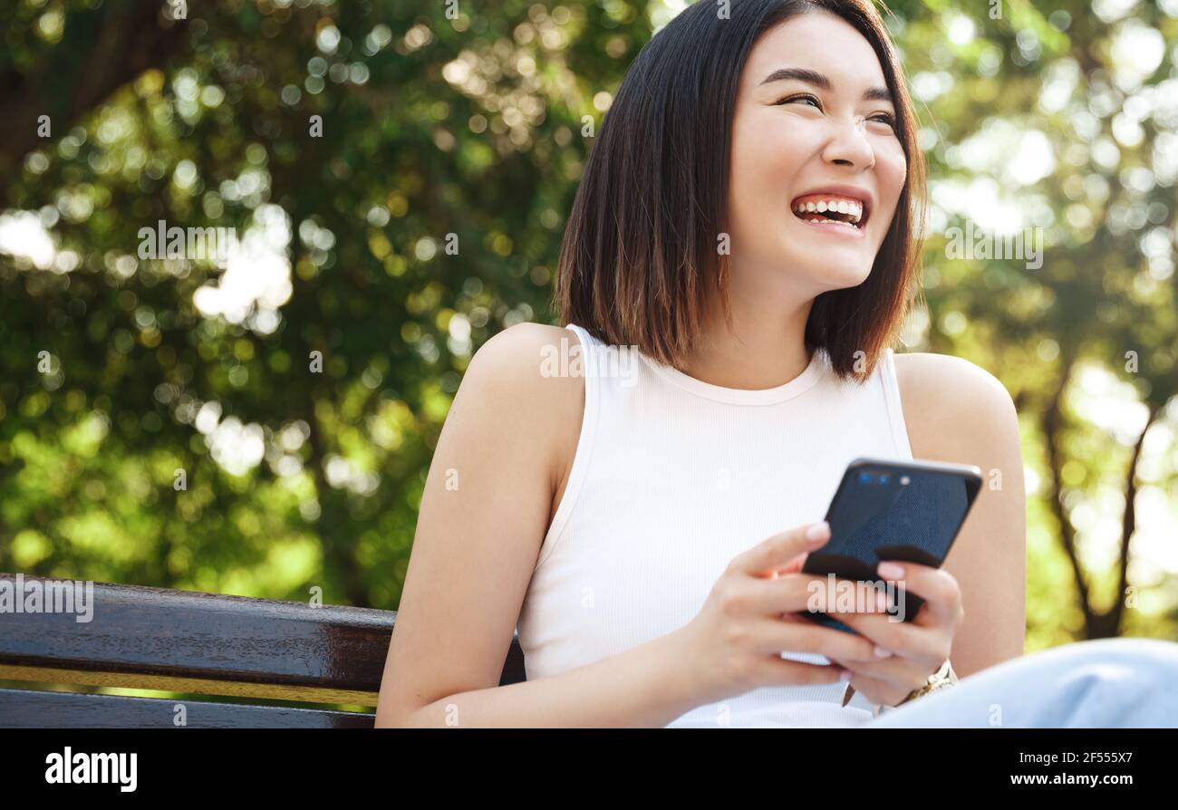 Glücklich asiatische Mädchen auf Bank sitzen und mit Handy. Mädchen, die sich im Park ausruhen und in sozialen Medien auf dem Smartphone surfen und jemanden anlachen Stockfoto