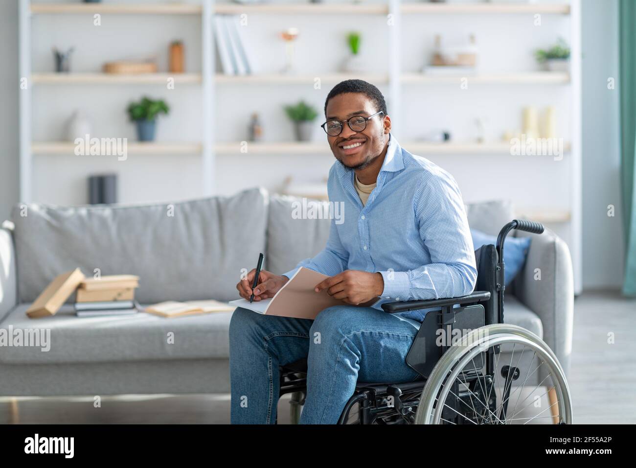 Glücklicher behinderter schwarzer Kerl im Rollstuhl, der Notizen macht, die Kamera anschaut und zu Hause lächelt, Kopierraum Stockfoto