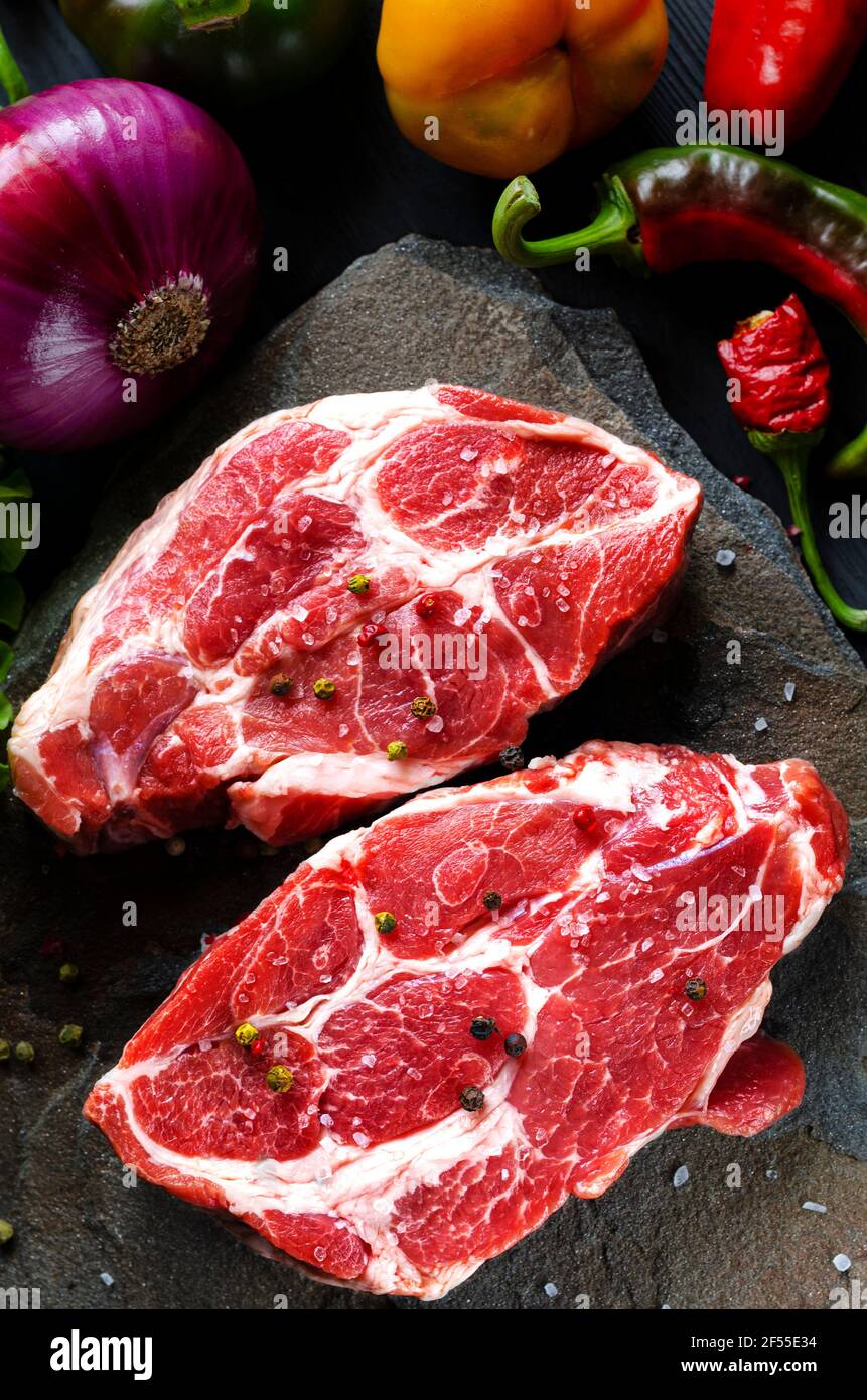 Fleisch rohe Steaks liegen auf schwarzem Hintergrund mit Gemüse, frischen blauen Zwiebeln, gelben Paprika, Chili. Hintergrundbild. Seitenansicht, Draufsicht Stockfoto