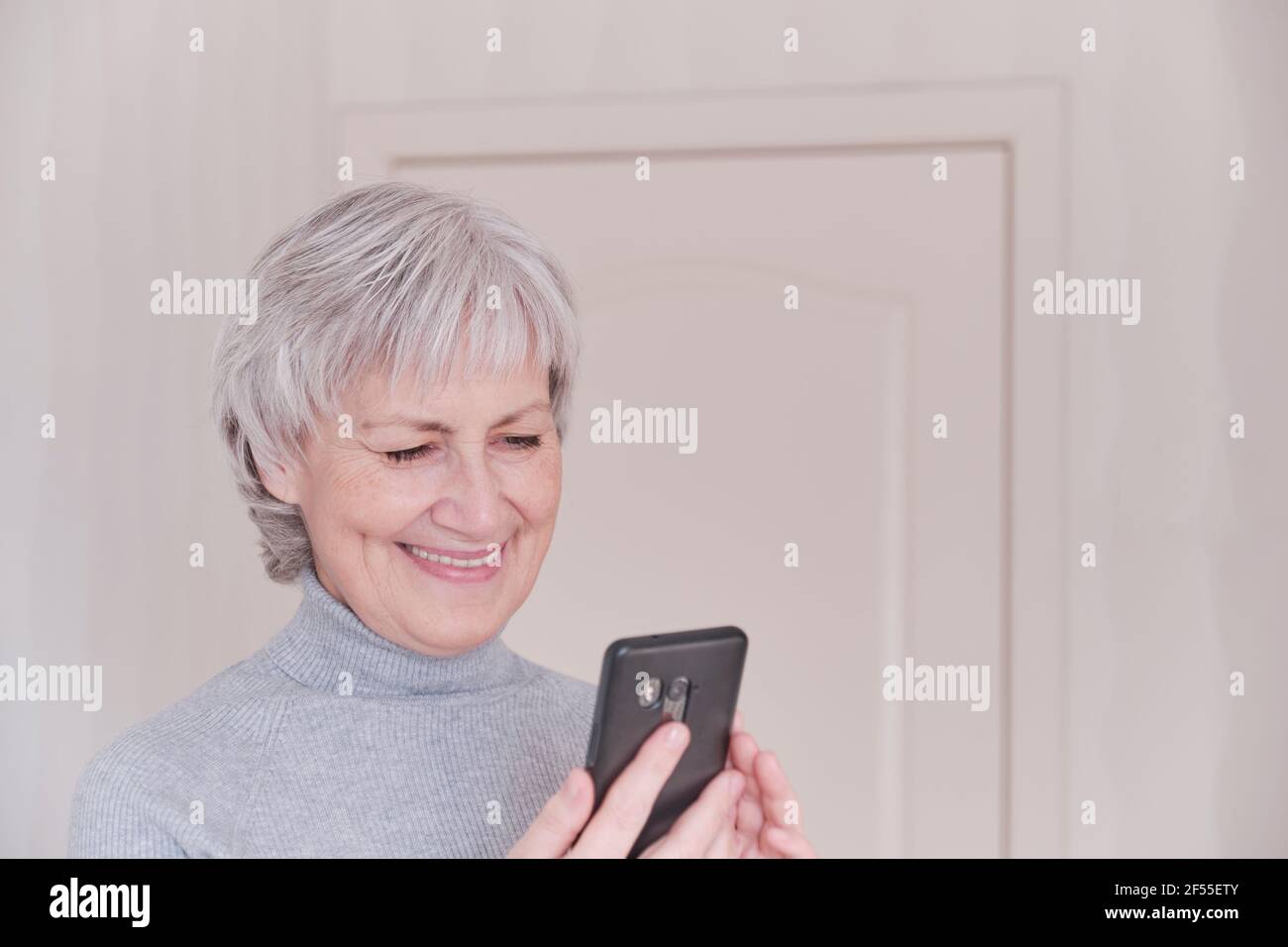 Zu Hause grauhaarige ältere Frau in grau Rollkragen schaut auf Smartphone , lächelnd. Menschen verbinden, Kommunikation. Lifestyle-Moment. Nahaufnahme Stockfoto
