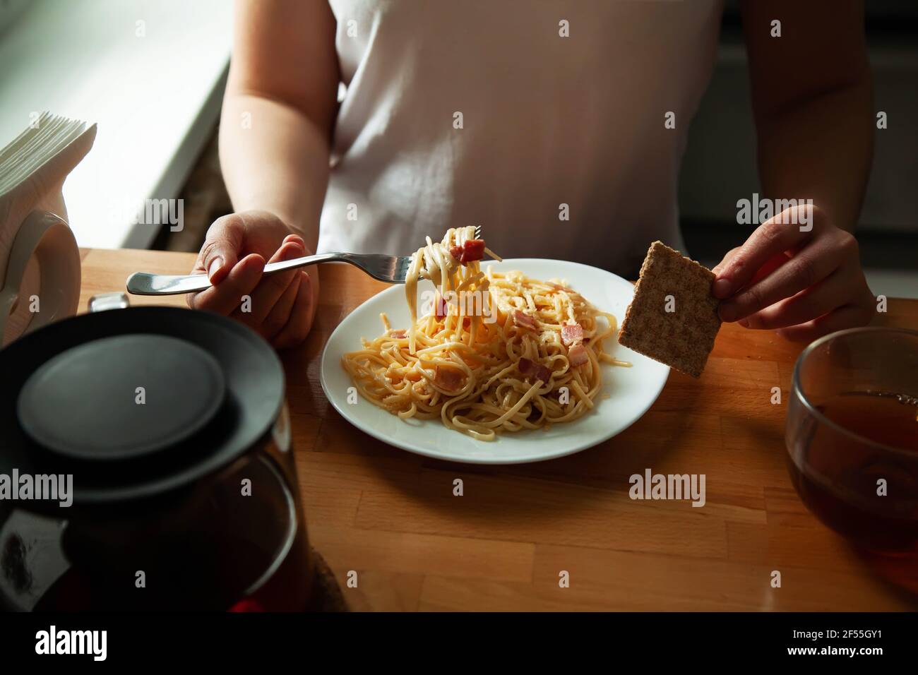 Ein Mädchen in hausgemachten Kleidern essen italienische Spaghetti, Carbonara am Tisch. Hausgemachtes Mittagessen, Hände mit einem Gericht Nahaufnahme Stockfoto