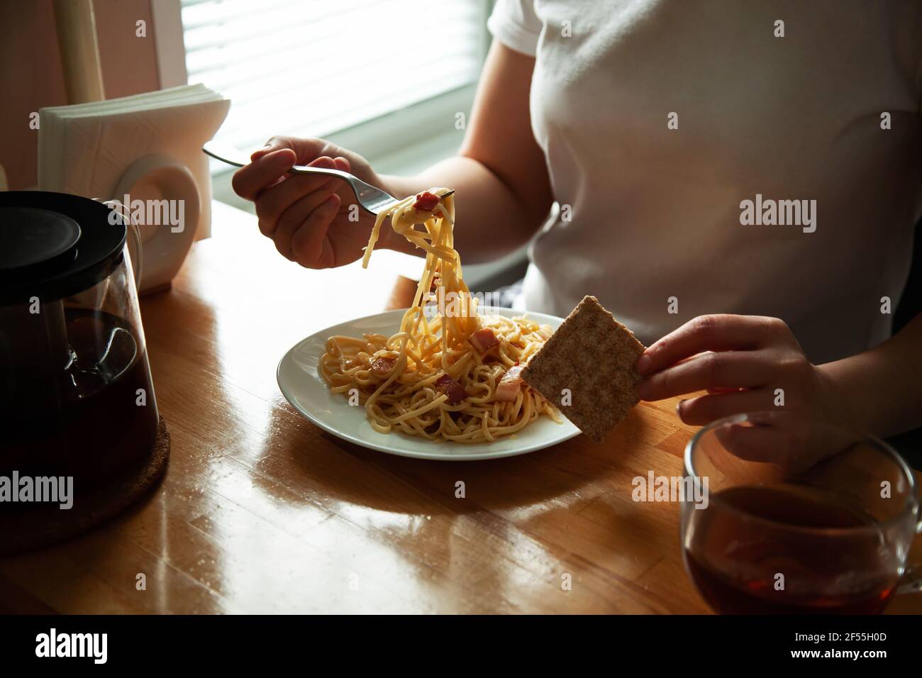 Ein Mädchen in hausgemachten Kleidern essen italienische Spaghetti, Carbonara am Tisch. Hausgemachtes Mittagessen, Hände mit einem Gericht Nahaufnahme Stockfoto