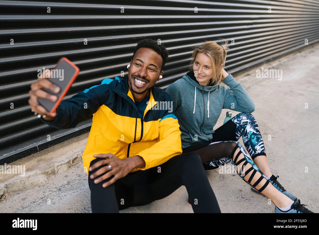 Fröhlicher Sportler mit Sportlerin, die während des Selfies über das Mobiltelefon unterwegs ist An der Wand sitzen Stockfoto