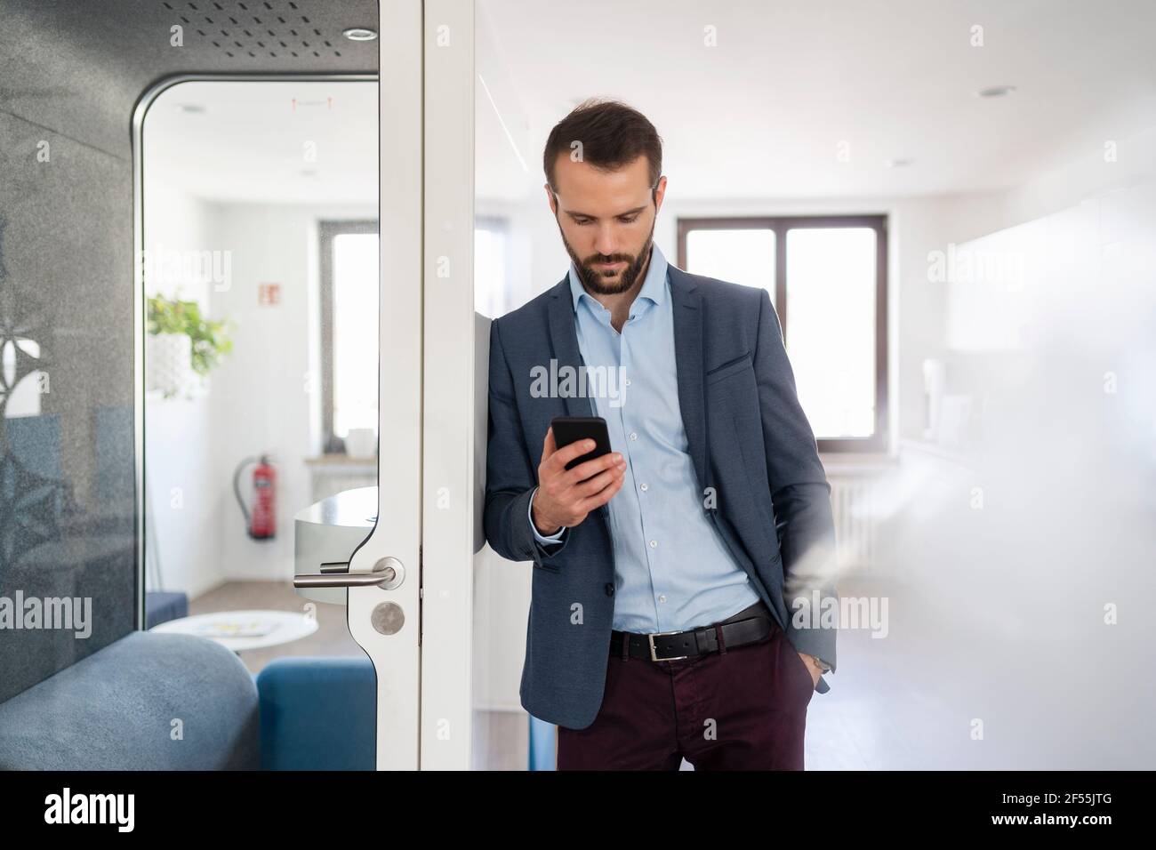 Junger Unternehmer, der Mobiltelefon verwendet, während er sich an der Telefonzelle stützt Im Büro Stockfoto