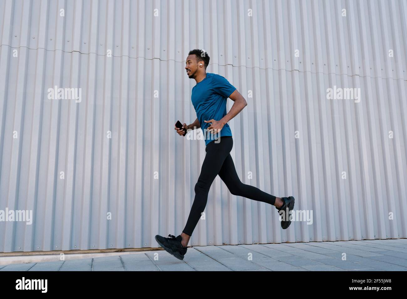 Männlicher Athlet mit Mobiltelefon, das an einer Wand auf dem Bürgersteig läuft Während des Trainings Stockfoto