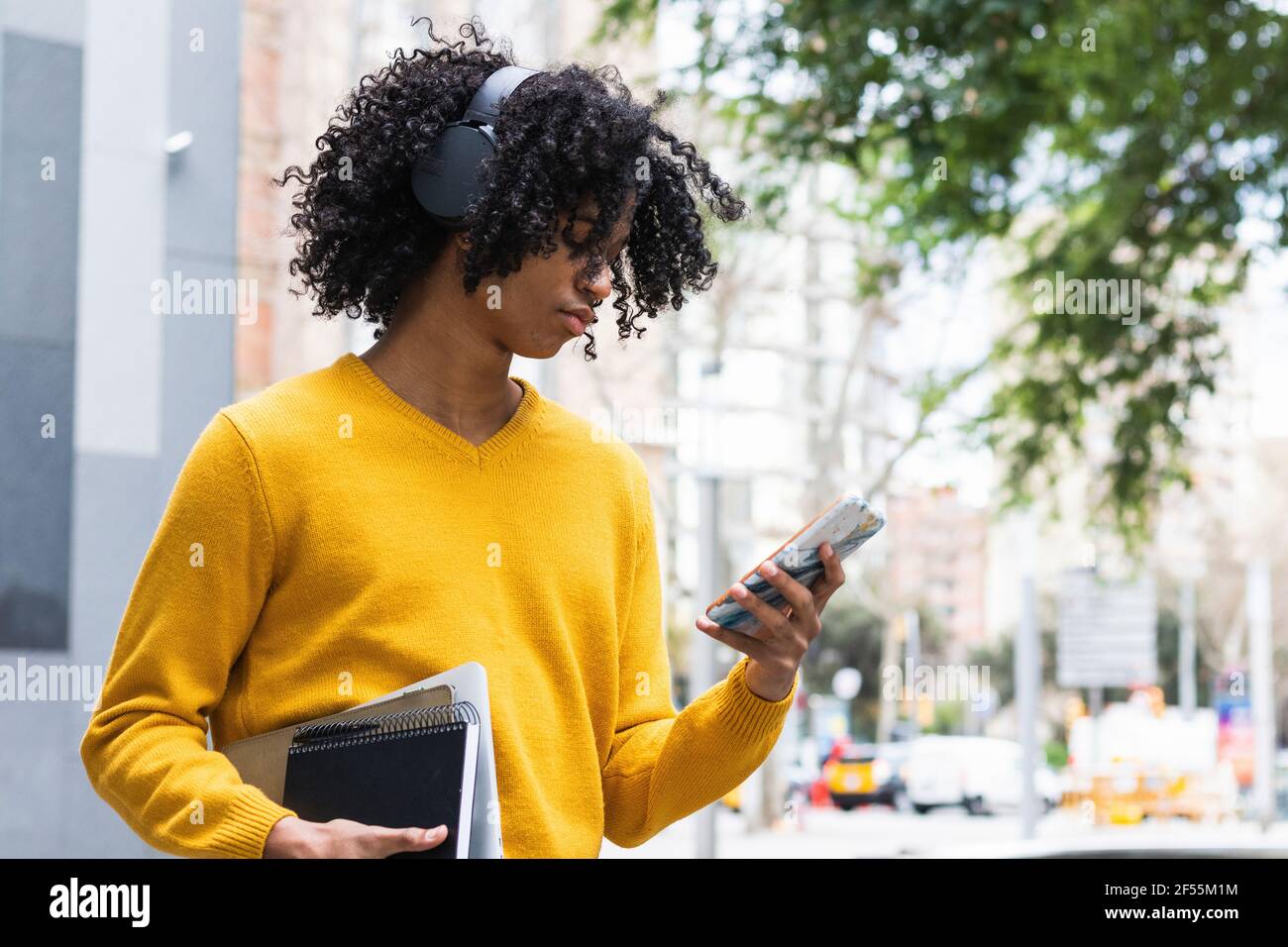 Junge, der Kopfhörer trägt und dabei mit einem Mobiltelefon steht, während er mit Büchern steht Im Freien Stockfoto