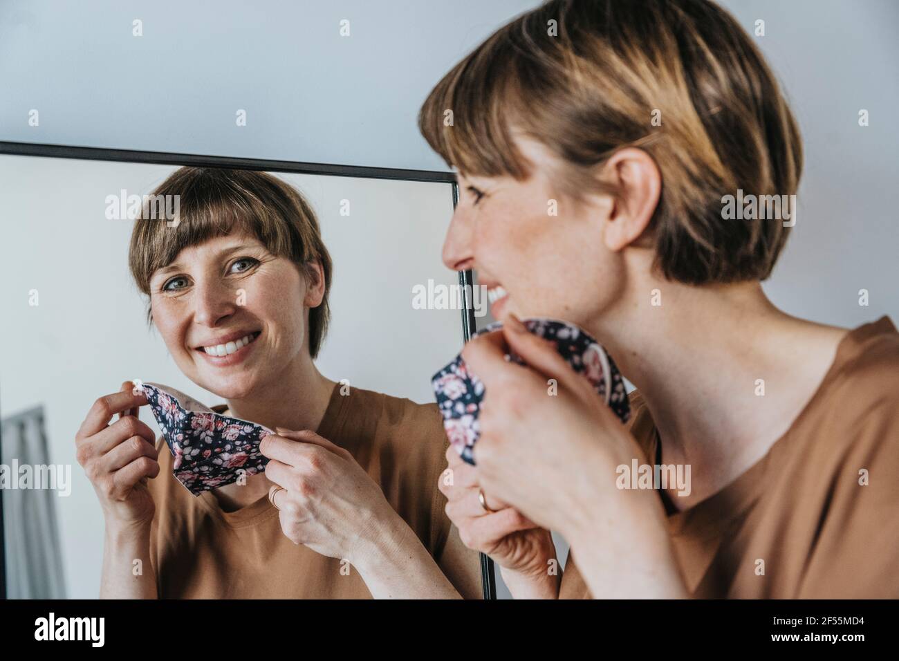 Glückliche Frau, die in den Spiegel schaut, während sie die schützende Gesichtsmaske hält Während einer Pandemie Stockfoto