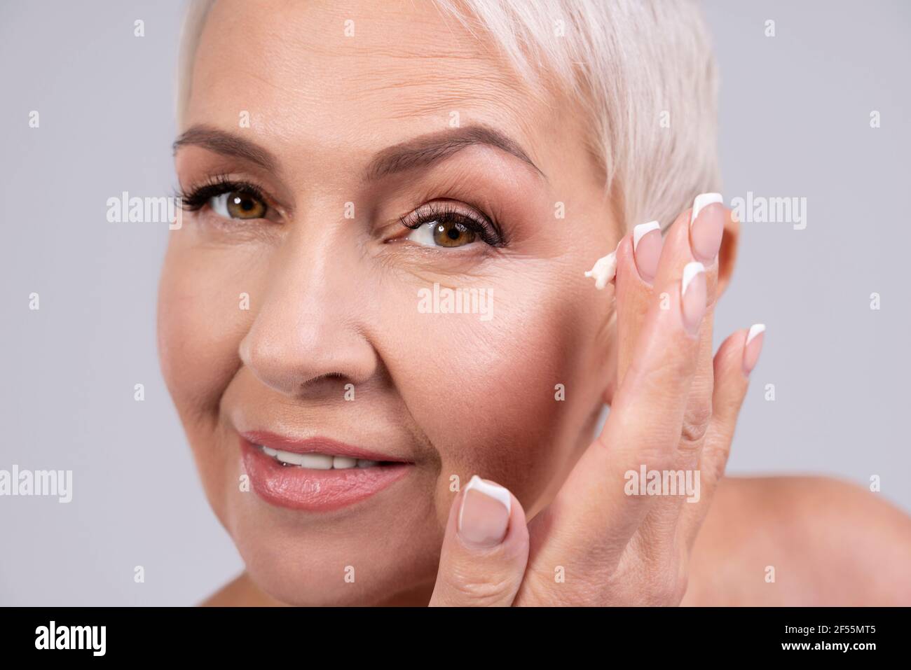 Lächelnde Frau, die Gesichtscreme von Falten auf grauem Hintergrund auftragen Stockfoto