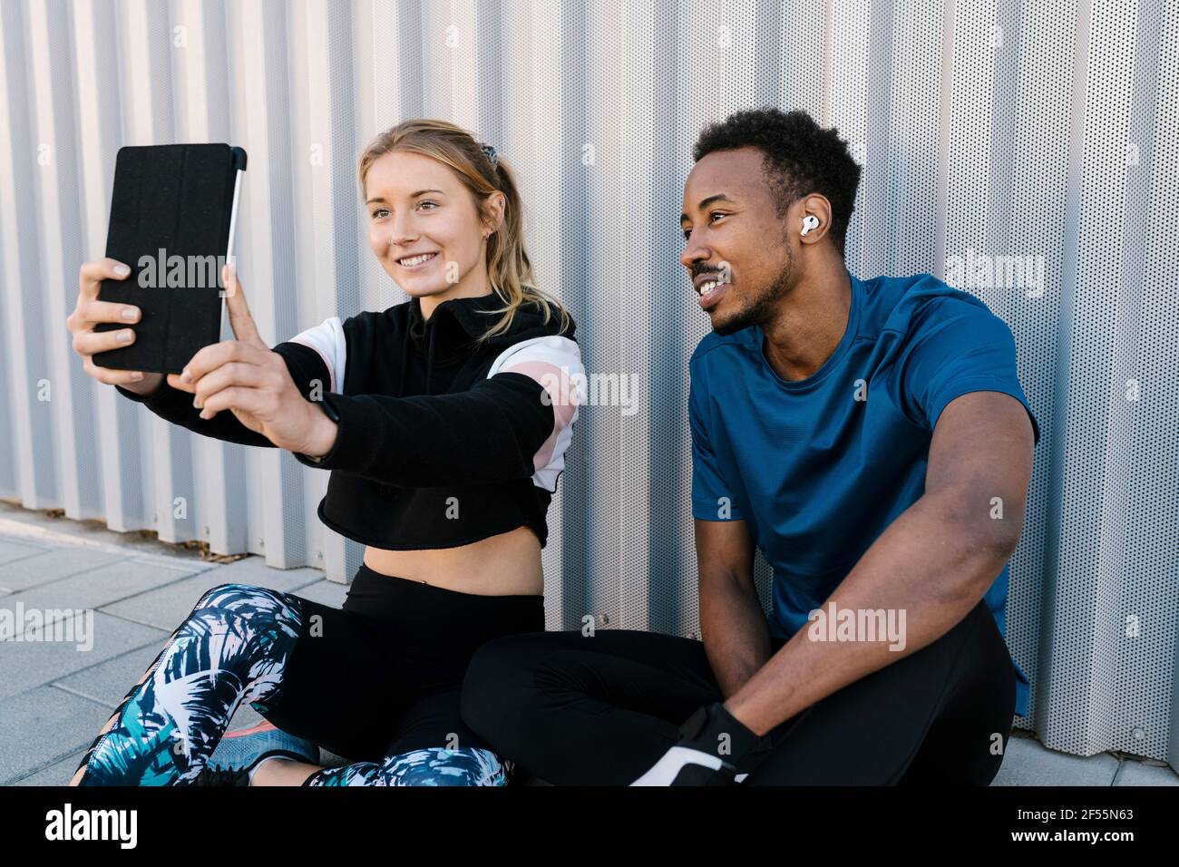 Junge Frau mit Mann nimmt Selfie durch digitale Tablette während Sitzen auf dem Boden gegen die Wand Stockfoto