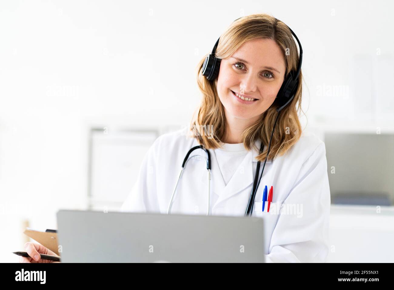 Lächelnde blonde Ärztin mit Kopfhörern während der Online-Konsultation bei Medizinische Klinik Stockfoto