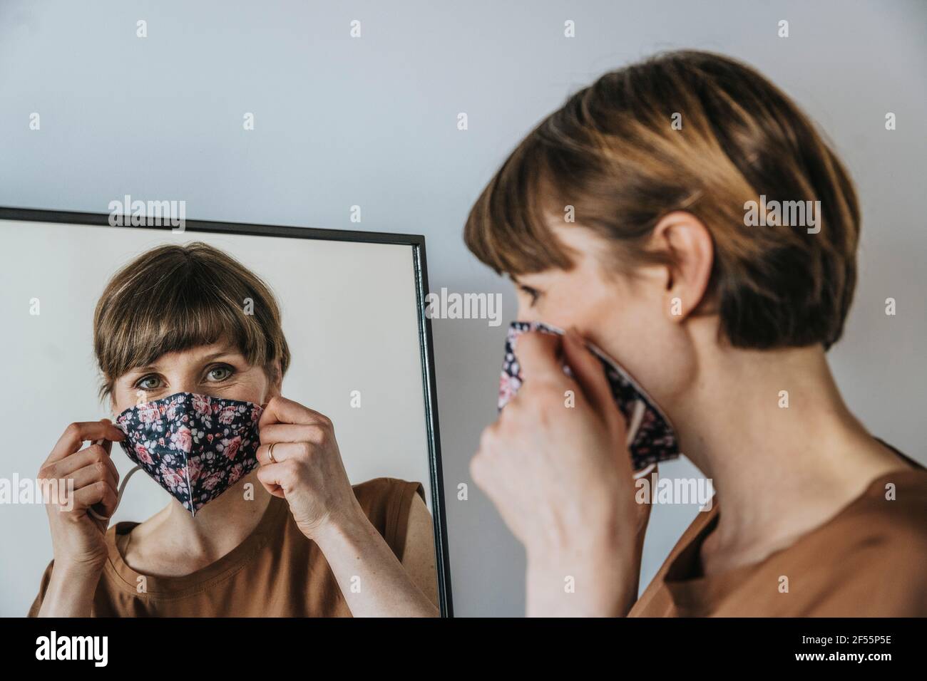 Braunhaarige Frau, die beim Blick ins Gesicht eine schützende Gesichtsmaske trägt Spiegel während einer Pandemie Stockfoto