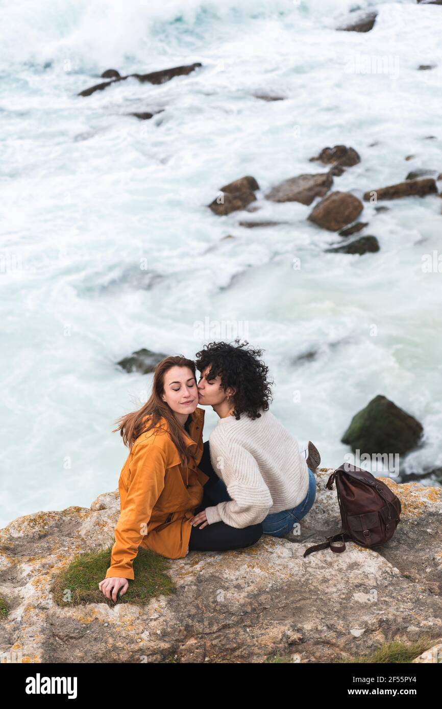 Junge Frau küsst die Wange eines Freundes, während sie auf einer Klippe sitzt Auf dem Seeweg Stockfoto