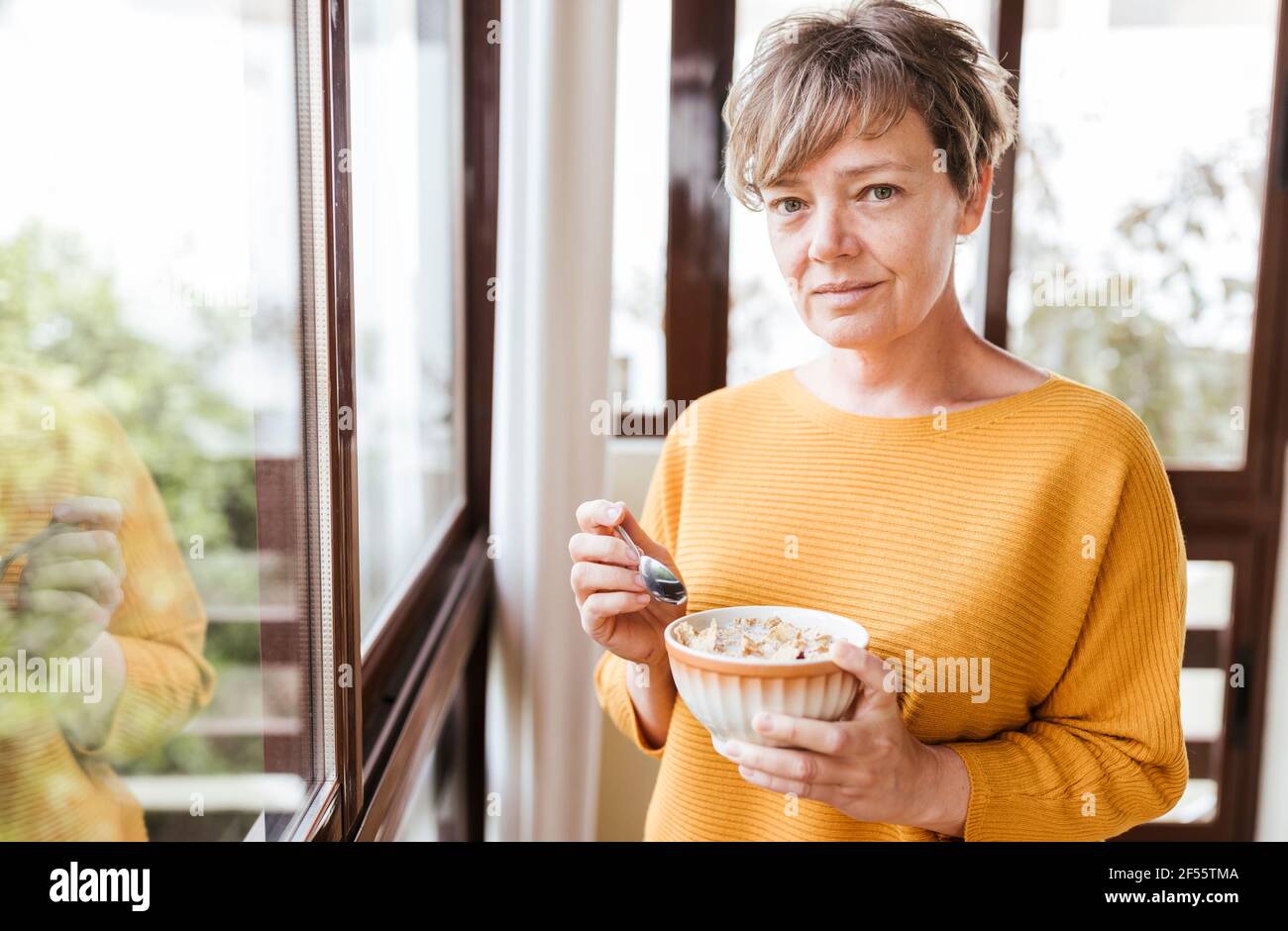 Frau mit kurzen Haaren beim Frühstück, während sie am Glasfenster steht Stockfoto