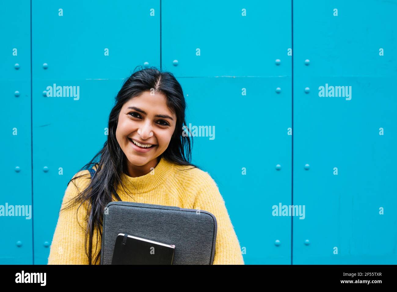 Glückliche Frau mit digitalem Tablet und Laptoptasche gegen Blau Wand Stockfoto