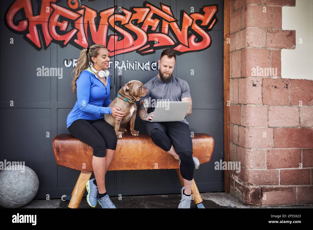 Männliche und weibliche Sportlerin, die einen Laptop benutzt, während sie mit einer Bulldogge sitzt Auf der Bank gegen die Graffiti-Tür Stockfoto