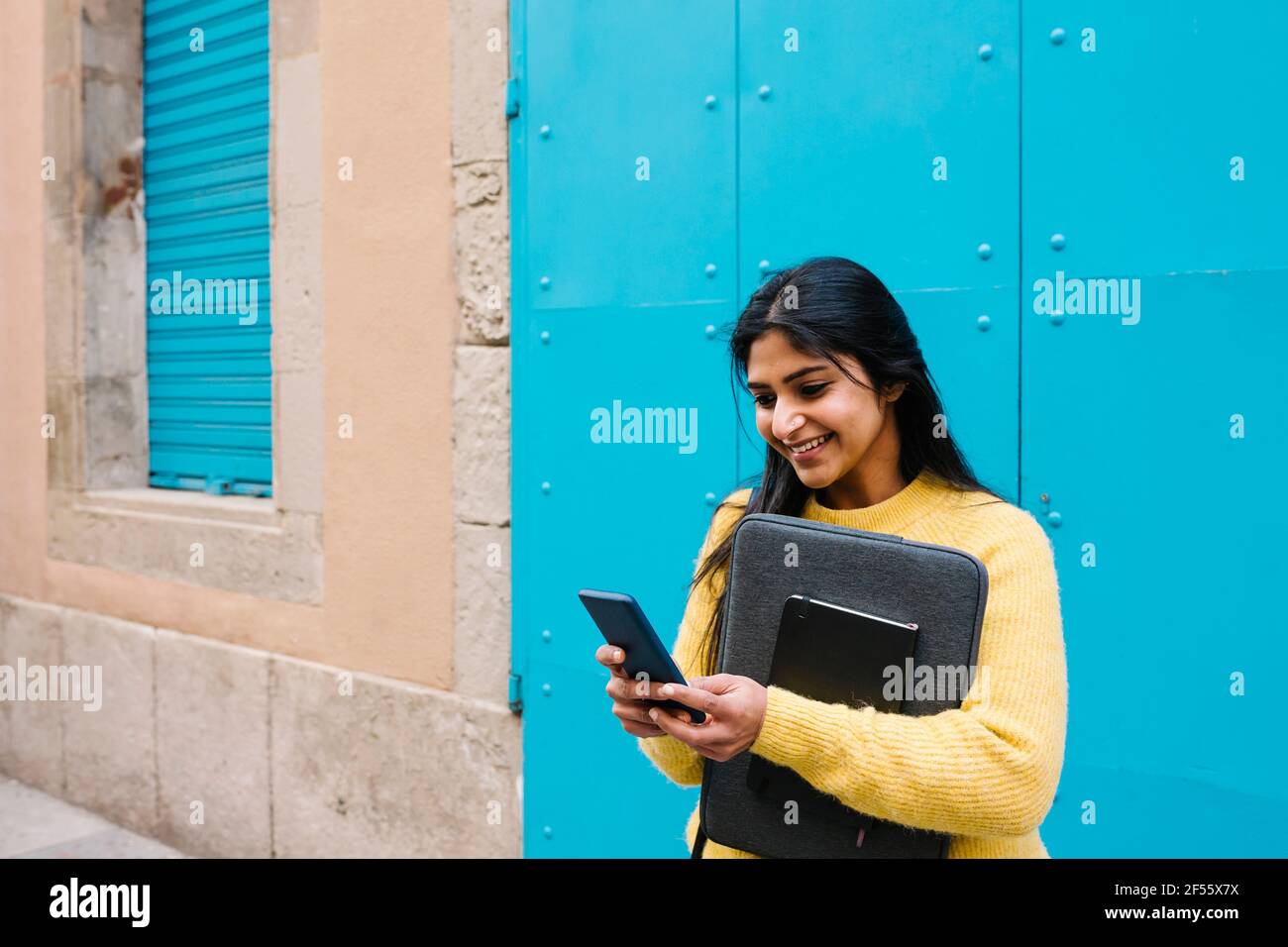 Lächelnde Frau mit digitalem Tablet und Laptoptasche mit Handy Telefon gegen blaue Wand Stockfoto