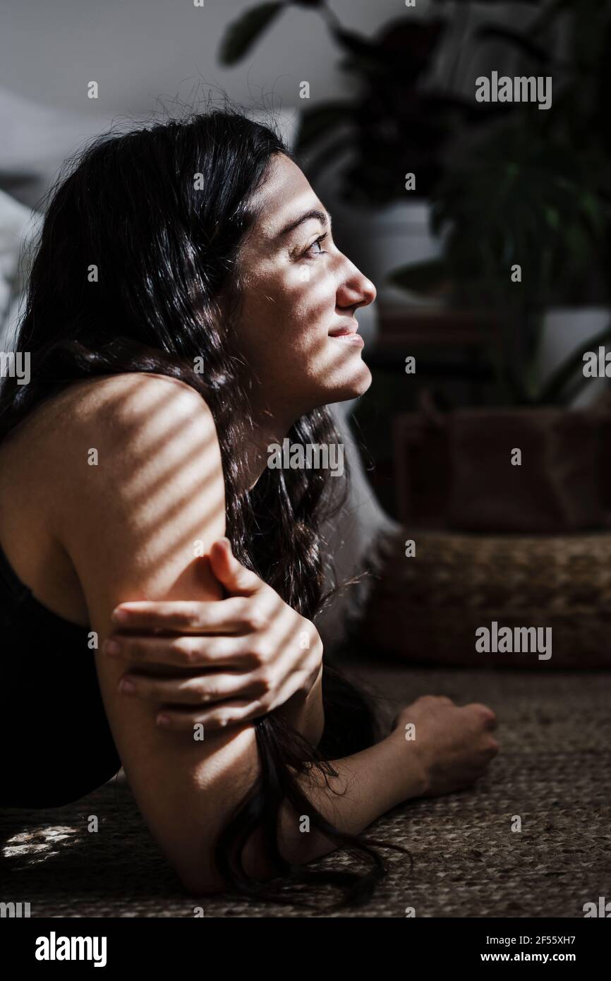 Lächelnde Frau mit Jalousien Schatten auf Gesicht wegschauen, während Liegen im Wohnzimmer zu Hause Stockfoto