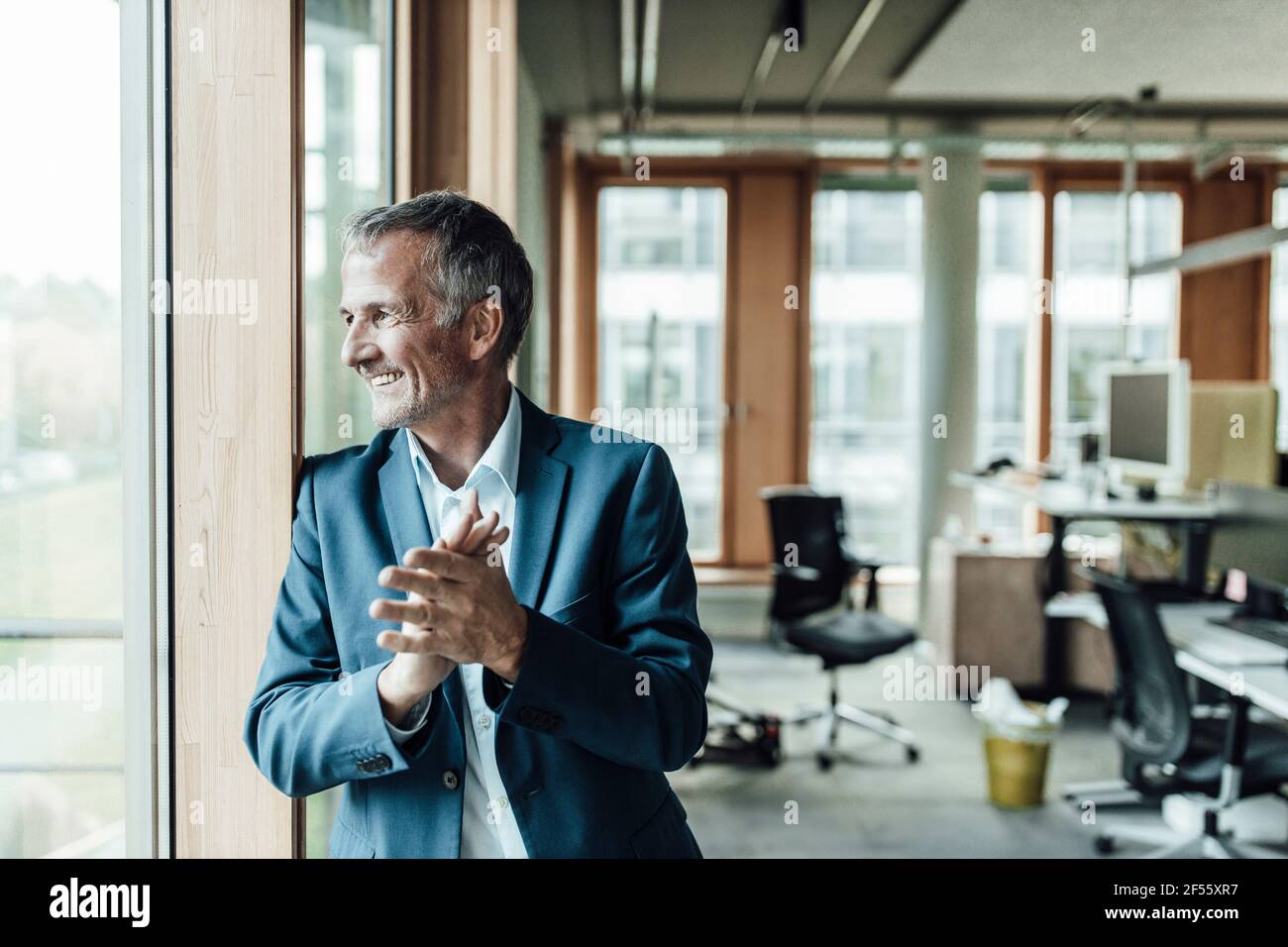 Lächelnder männlicher Unternehmer, die Hände zusammengeklemmt und durch das Fenster geschaut Büro Stockfoto
