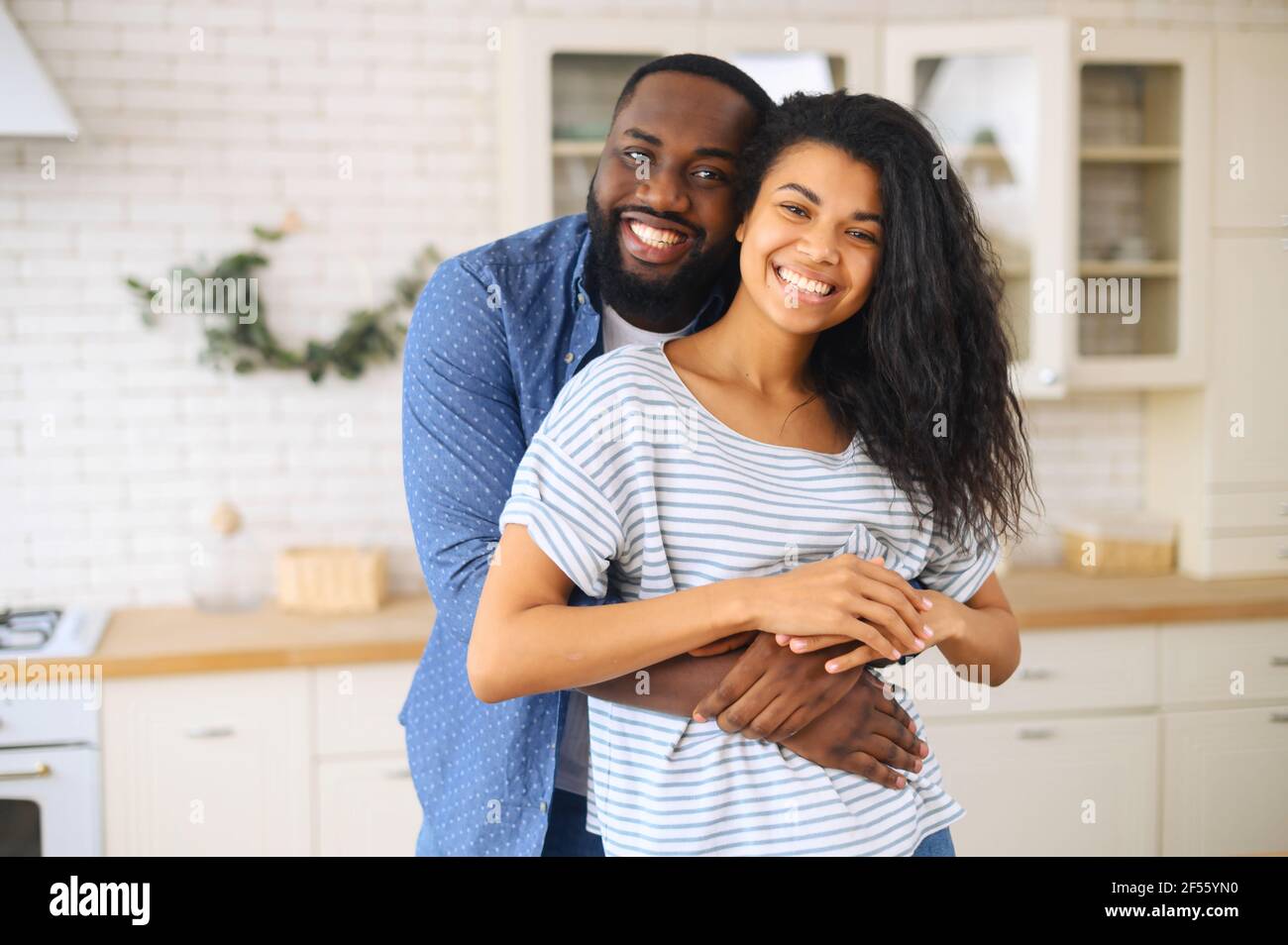 Glücklich verheiratet multirassischen Paar posiert für ein Foto in der Küche, die glücklichsten Herzen machen die glücklichsten Häuser, afrikanischen Mann umarmt lateinische Frau mit dicken lockigen Haaren von hinten, harmonische Beziehungen Stockfoto