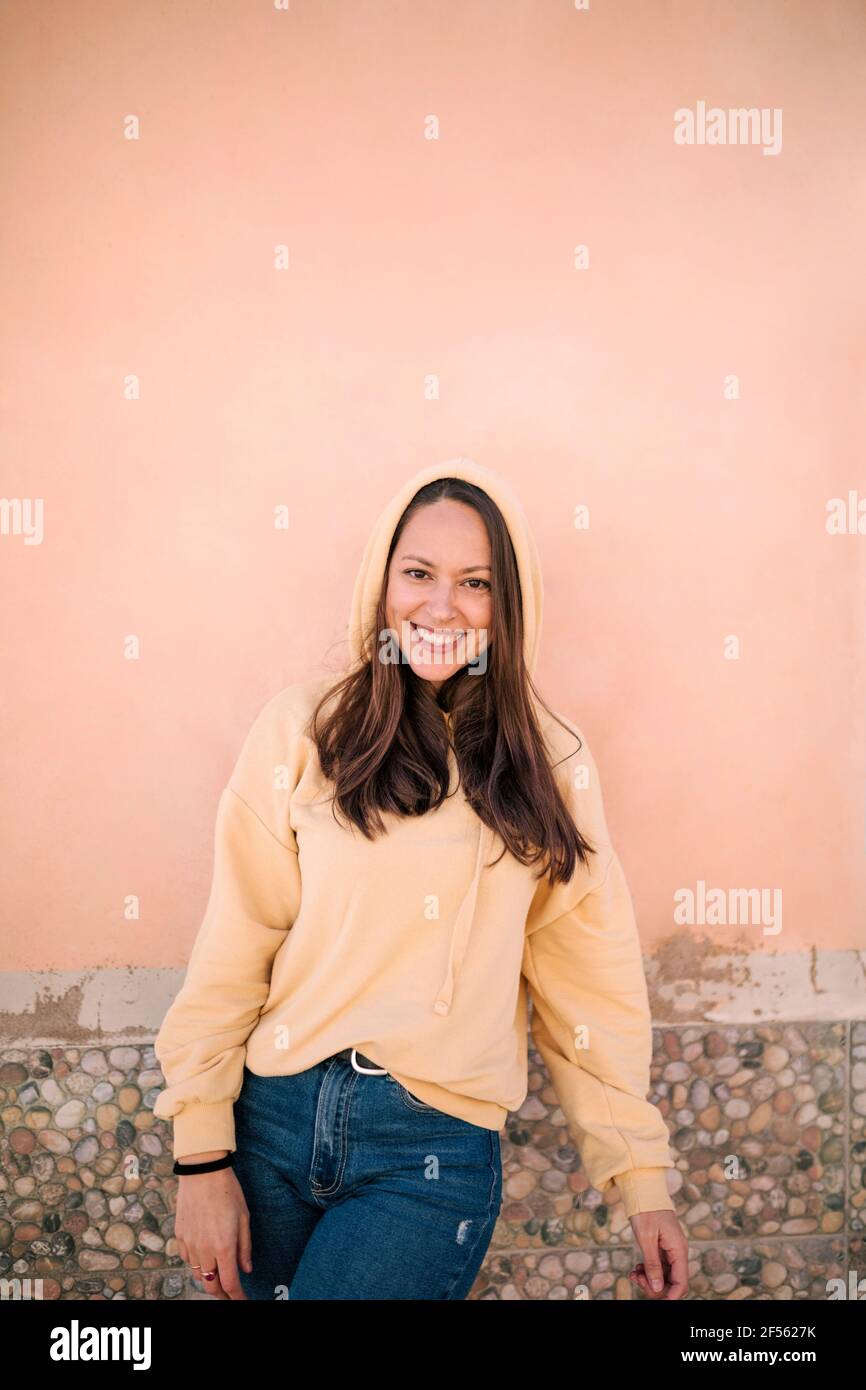 Schöne lächelnde weibliche Brünette trägt gelben Hoodie gegen rosa Wand Stockfoto