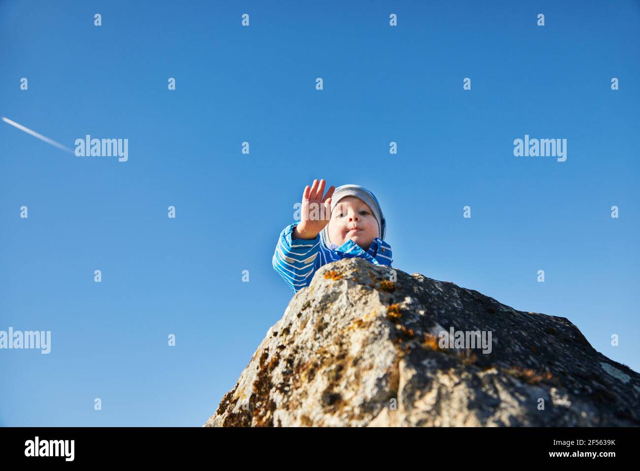 Niedlicher Junge auf Felsen, der die Hand gegen den Himmel winkt Stockfoto