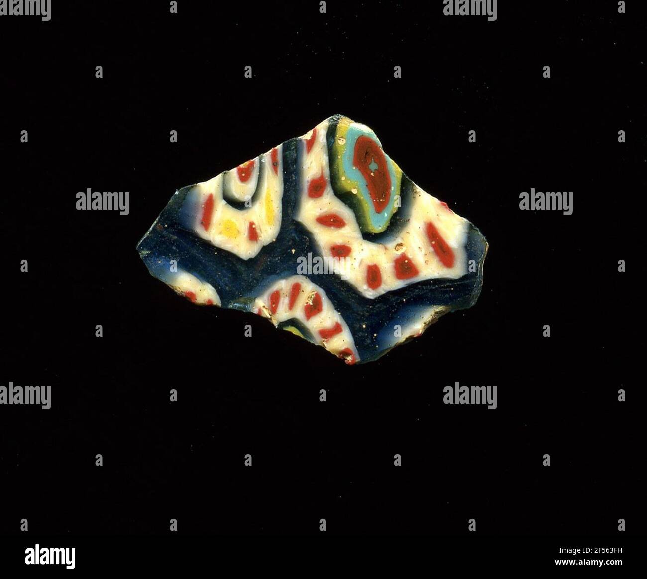 Mosaikglasfragment. Das unregelmäßig gebrochene Fragment hat millefiori-Blüten auf blauem Grund; diese bestehen aus roten Punkten mit weißem Rand zu Kreis, angeordnet mit blauen, gelben, türkisfarbenen und roten Kreisen von außen; eine andere Art von Blumen mit abwechselnd gelben und roten Punkten mit weißem Rand mit blauen, weißen, roten Kreisen innen. In der Römerzeit verwendete man farbiges, ornamentes Glas für Gefäße, Mosaike, Wanddekorationen und Möbelornamente. Für die Herstellung von verschiedenfarbigen Gläsern zu Röhren und Stangen. Diese wurden komprimiert und erhitzt, was zu einem langen Balken führte. Das Glas wurde in Scheiben geschnitten Stockfoto