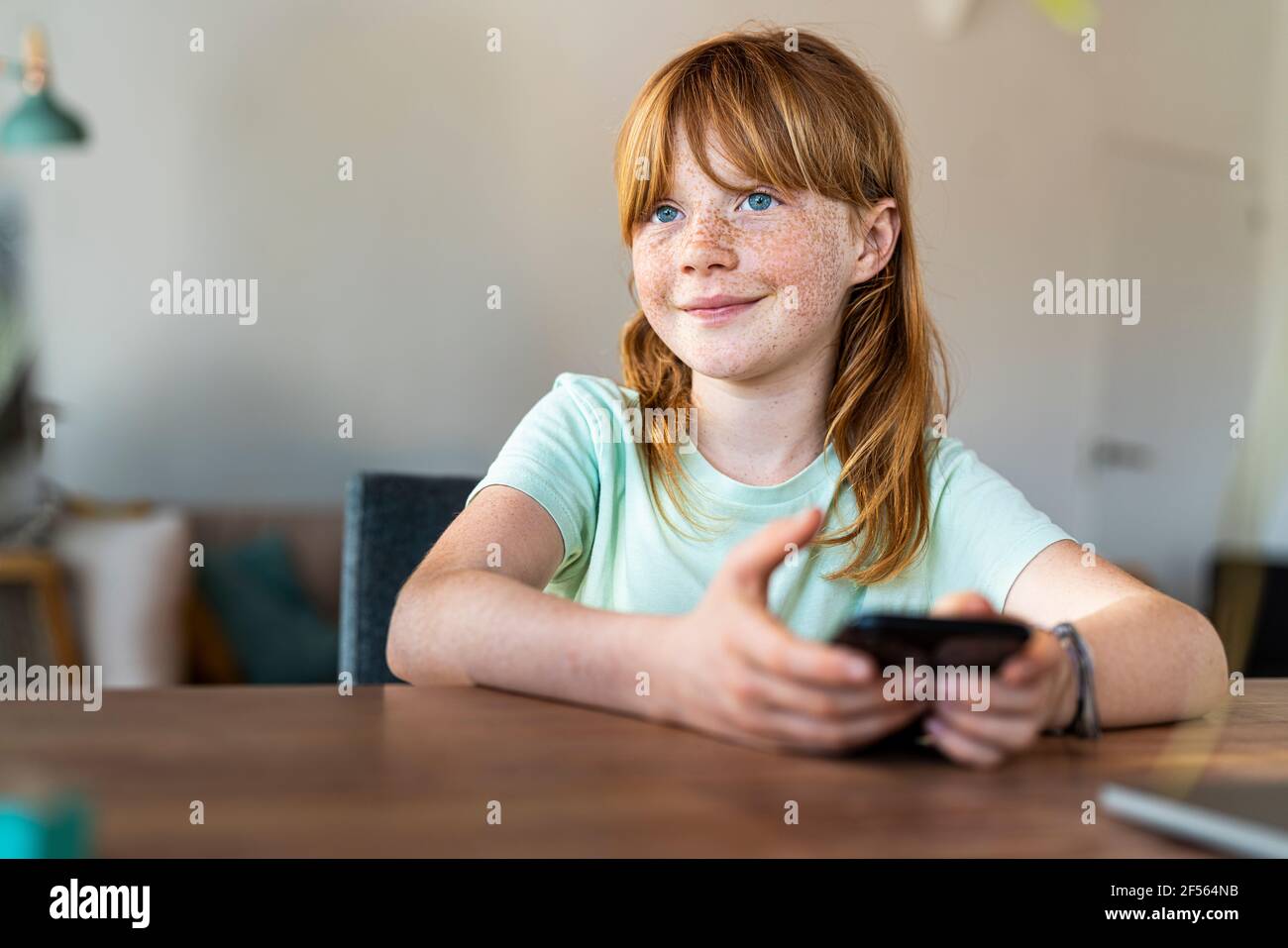 Nettes lächelndes Mädchen mit blauen Augen, das wegschaut, während es hält Smartphone zu Hause Stockfoto