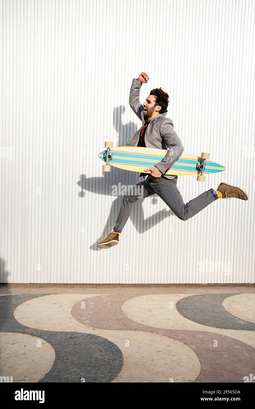 Fröhlicher Geschäftsmann mit Longboard, der an einer Wand auf einem Fußweg springt Stockfoto