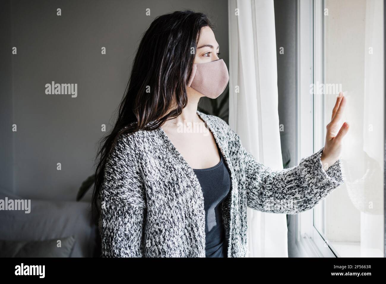 Frau mit schützender Gesichtsmaske, die im Stehen durch das Fenster schaut Im Wohnzimmer zu Hause während der COVID-19 Stockfoto