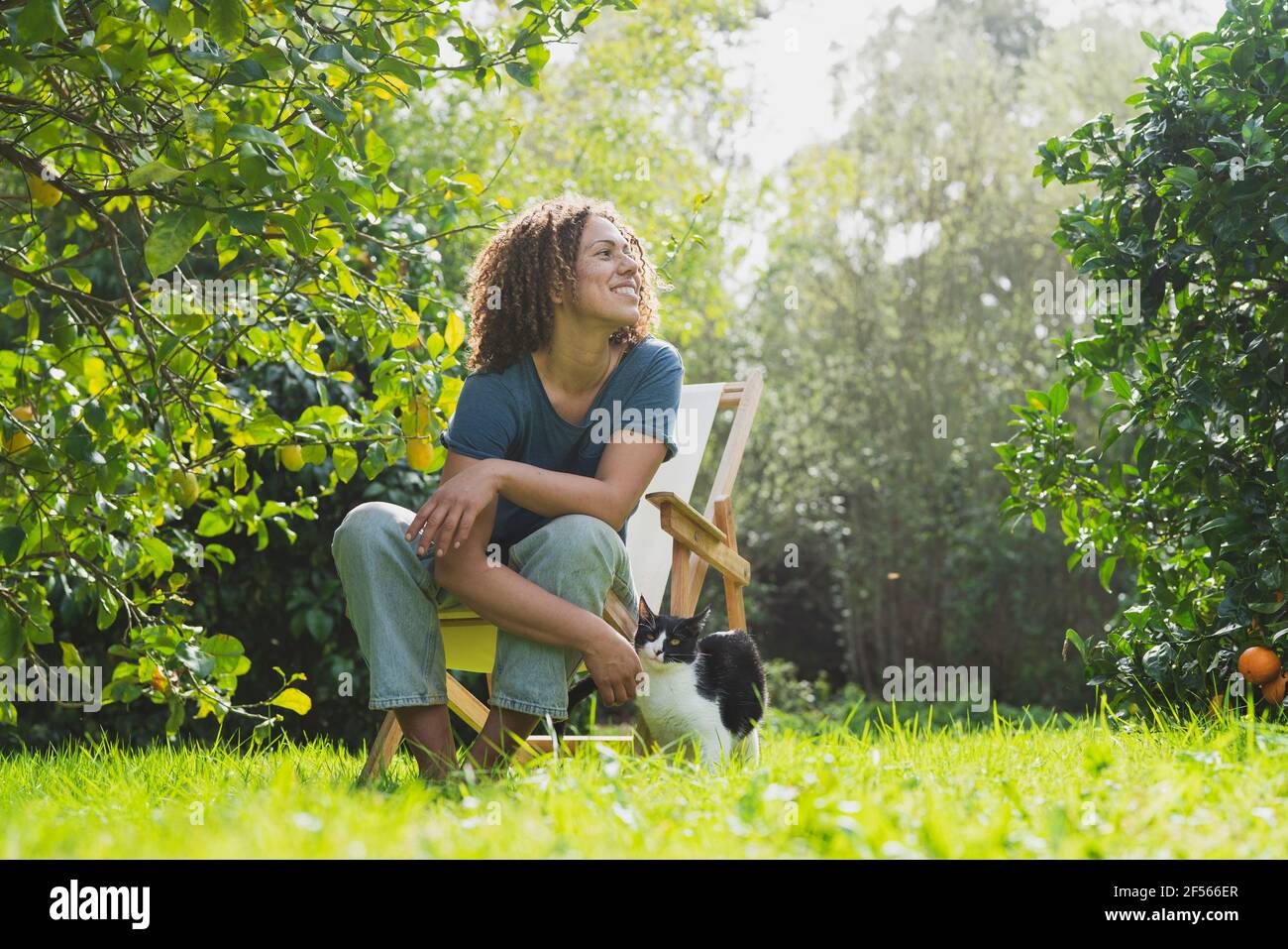 Lächelnde, lockige Frau schaut weg, während sie auf einem Stuhl sitzt Mit der Katze im Garten Stockfoto
