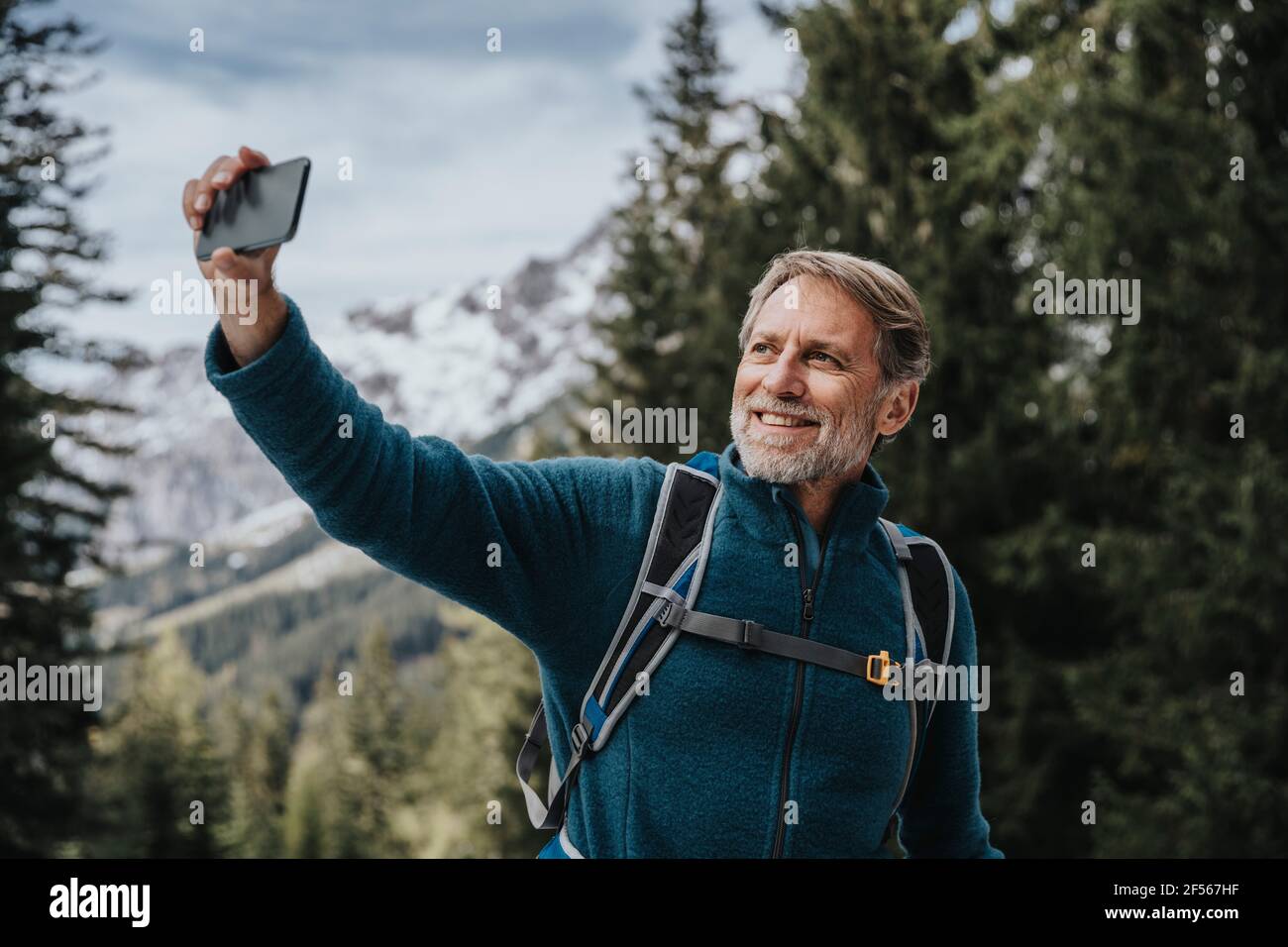 Lächelnder Mann, der im Salzburger Land, Österreich, Selfie per Smartphone gegen Baum macht Stockfoto