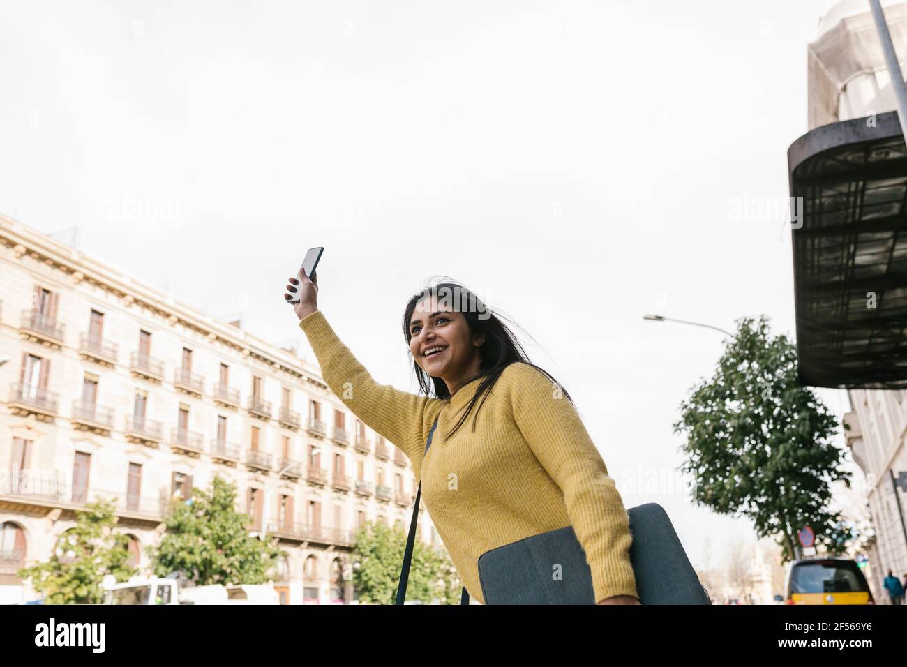 Lächelnde Frau mit Handy, die ein Taxi gegen den Himmel hagelt Stockfoto