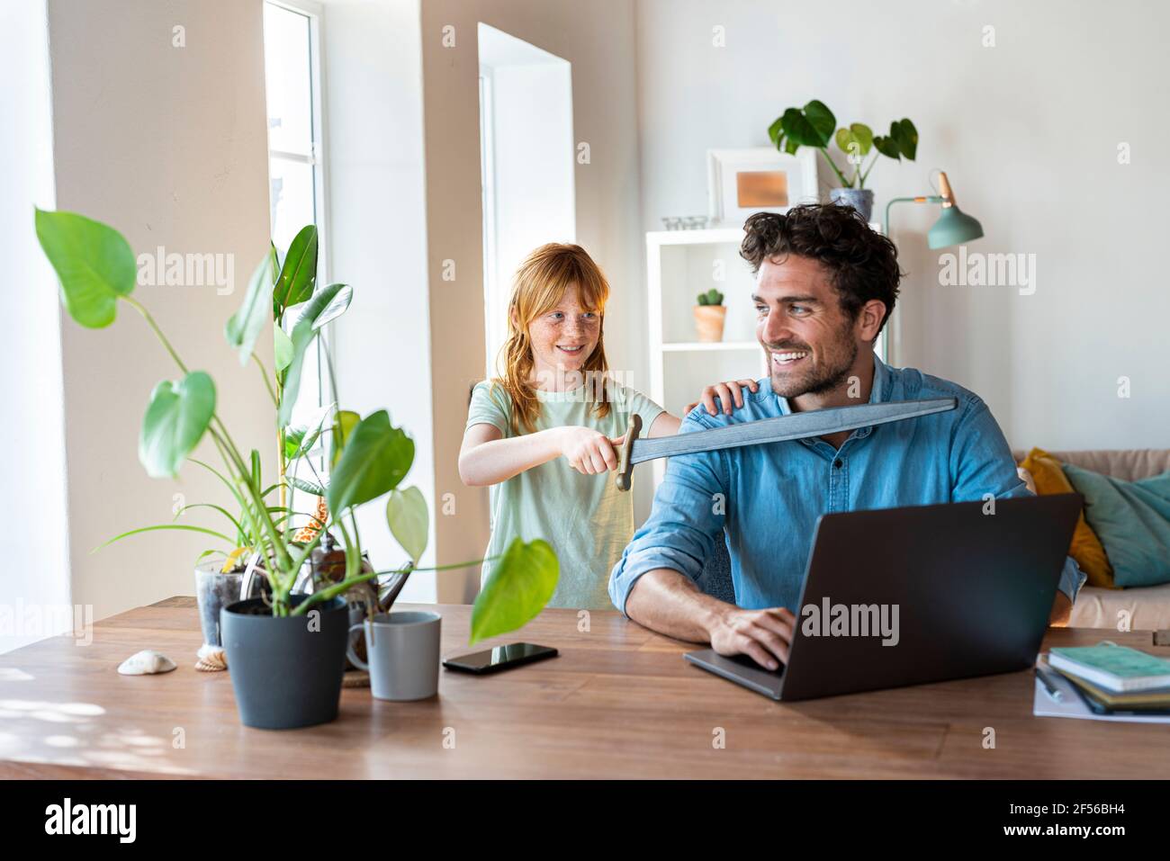 Verspielte Mädchen mit Spielzeug Schwert spielen mit Vater arbeiten an Laptop zu Hause Stockfoto