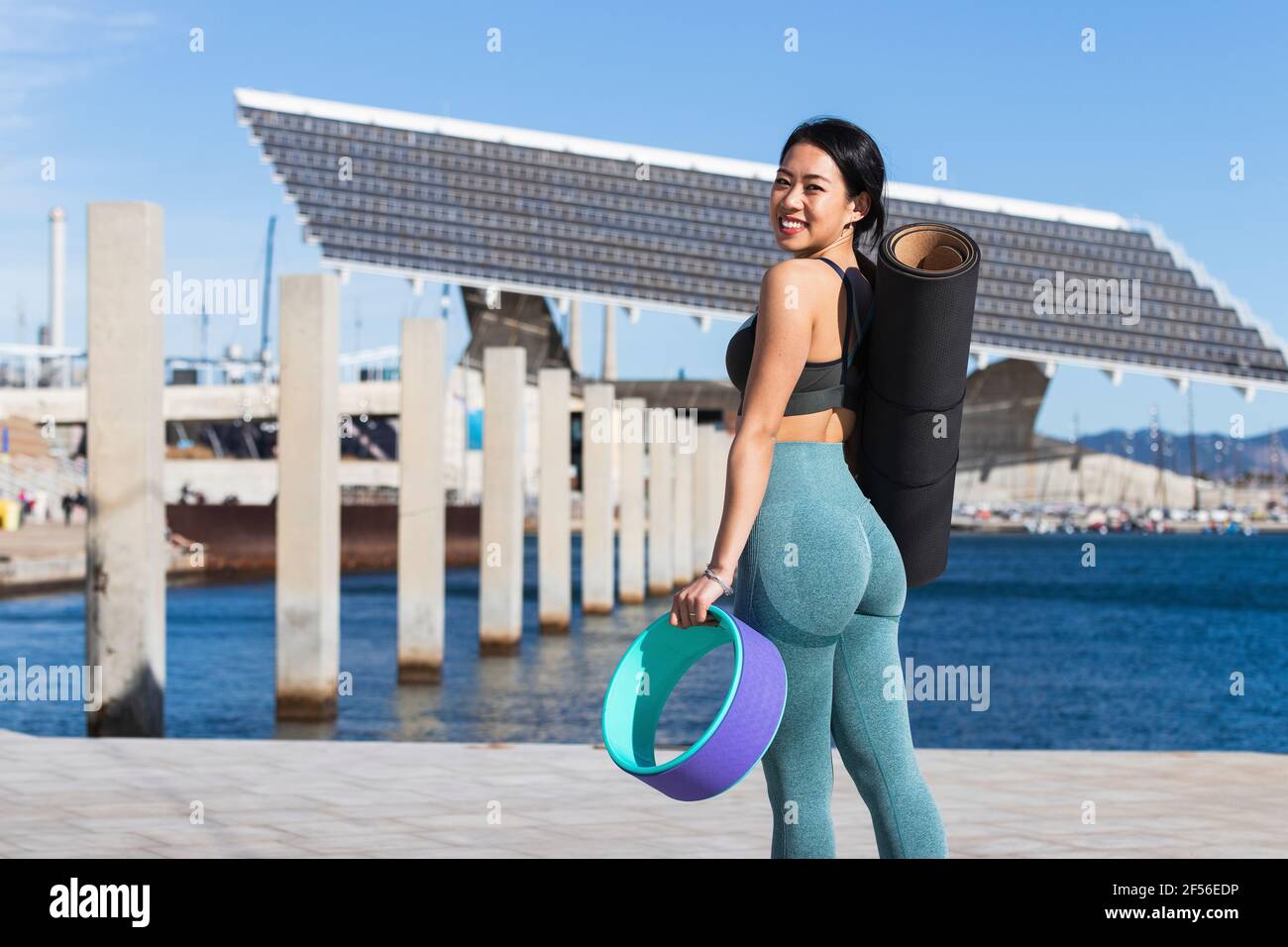 Glückliche Chinesin, die Yogamatte und Rad an der Promenade trägt Stockfoto